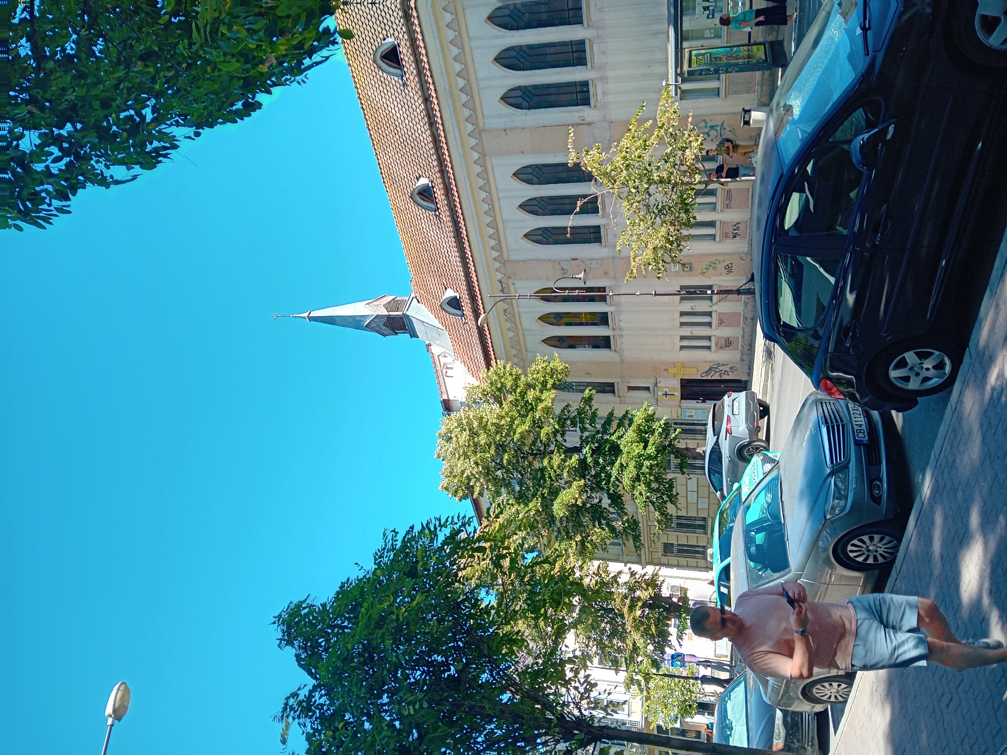 The image depicts a street scene in Varna, Bulgaria, under a clear blue sky. In the foreground, a man in a pink t-shirt and light blue shorts is walking on the sidewalk, looking at his phone. He is wearing sunglasses and a watch. Several cars are parked along the street, including a dark-colored Mercedes-Benz with a Bulgarian license plate "CB 4112XH" and a dark hatchback. Behind the cars, a building with a tiled roof and arched windows stands. A church steeple is visible above the building. Trees line the street, providing shade. In the background, there is a bus stop shelter. The overall impression is a sunny day in an urban environment.