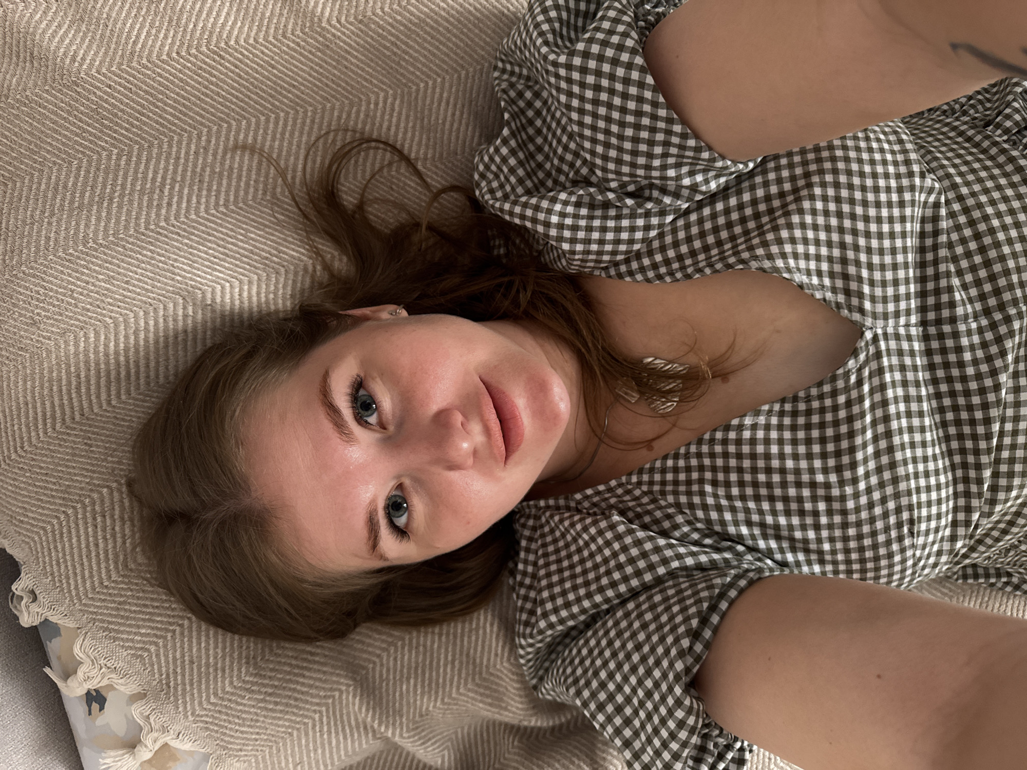 A woman with light brown hair and blue eyes is lying on a beige herringbone-patterned blanket or bedspread. She is wearing a green and white checkered dress. Her head is tilted slightly to the left. The background includes a section of a gray object with a floral or decorative pattern. No definitive connection to Ферма, Russia can be established based on the visual information provided.