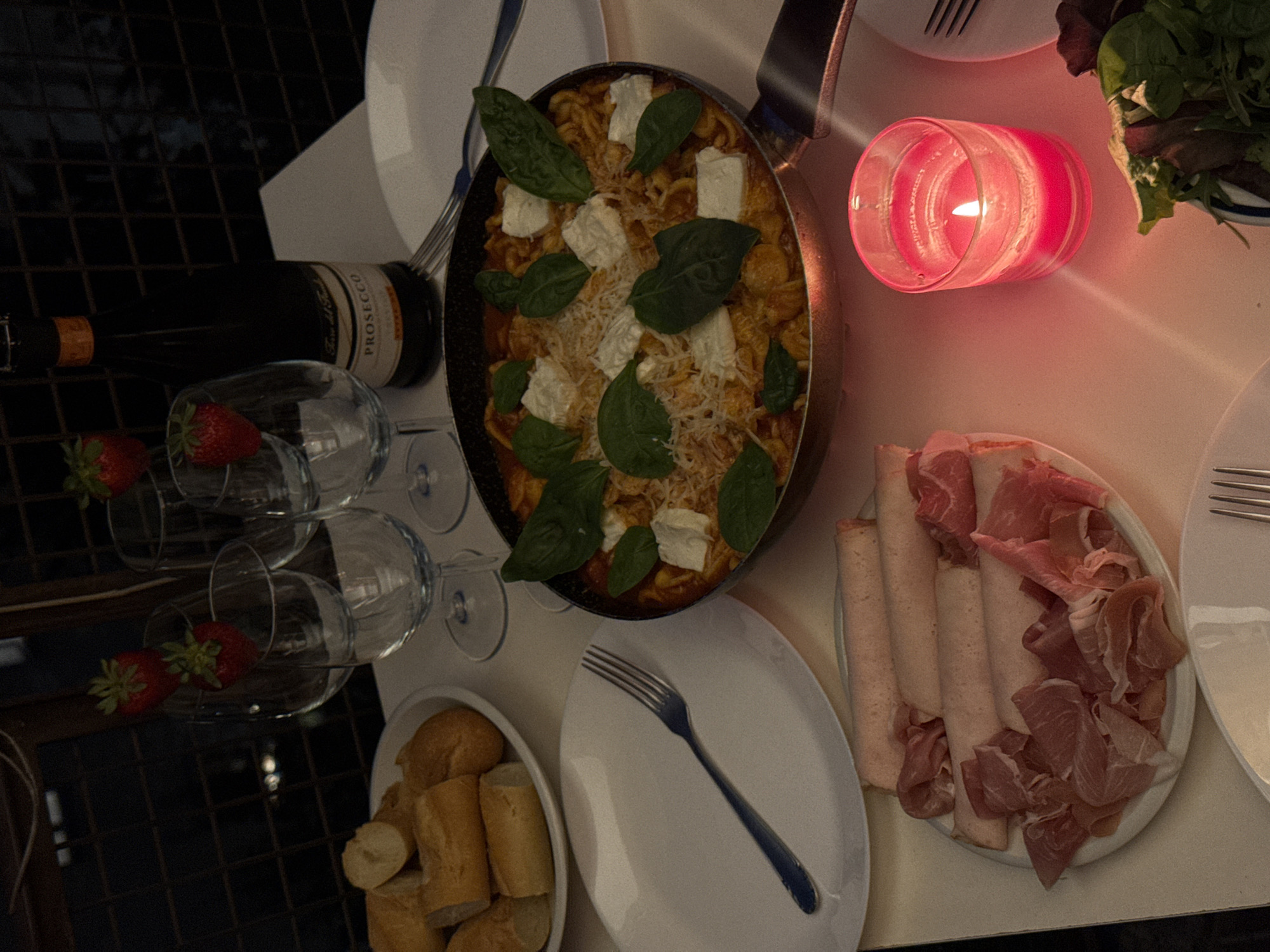 The image presents an overhead view of a table set for an evening meal, bathed in the warm, dim glow of a red candle. No people are visible, but the setup suggests an intimate dinner, possibly in Milan, Italy.
Central to the scene is a dark pan filled with a pasta dish, featuring small pasta shells in a red sauce. It is generously garnished with fresh green basil leaves, dollops of white cheese (likely ricotta or mozzarella), and grated cheese. To the left, a dark bottle of "PROSECCO" rests beside several clear stemmed glasses. Two of these glasses each hold a single red strawberry, ready for a drink.
To the bottom left, a white bowl holds an assortment of bread rolls. On the middle-right, a white oval plate displays a selection of cured meats, including thinly sliced prosciutto and rolled cuts of what appears to be turkey or ham. A smaller bowl in the upper right corner contains a green salad. A white plate with a dark fork is also visible, indicating a place setting.
The background is dark with a subtle grid pattern, suggesting a floor or dark surface in a dimly lit indoor or sheltered outdoor setting. The overall ambiance is cozy and inviting, emphasizing the prepared food and drink for a relaxed dining experience.