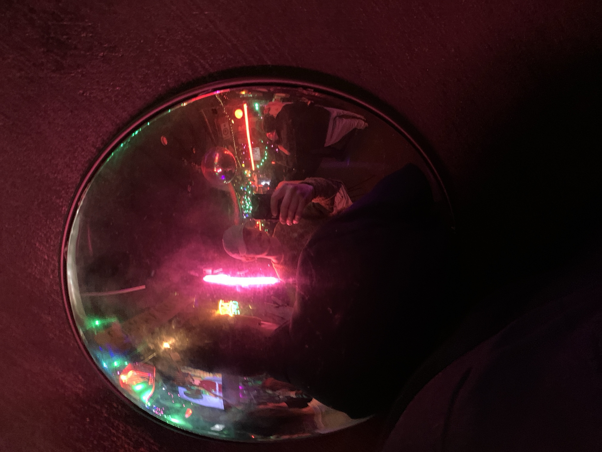 A person is taking a photo through a porthole-style window or opening. The scene inside the window is illuminated with colorful lights, including what appears to be a disco ball and neon signs. The person taking the photo is partially visible on the right side, wearing dark clothing. The environment surrounding the window is dark and appears to be inside a tunnel or enclosed space with textured, maroon-colored walls. There is no explicit information about the location in Tel-Aviv, Israel.