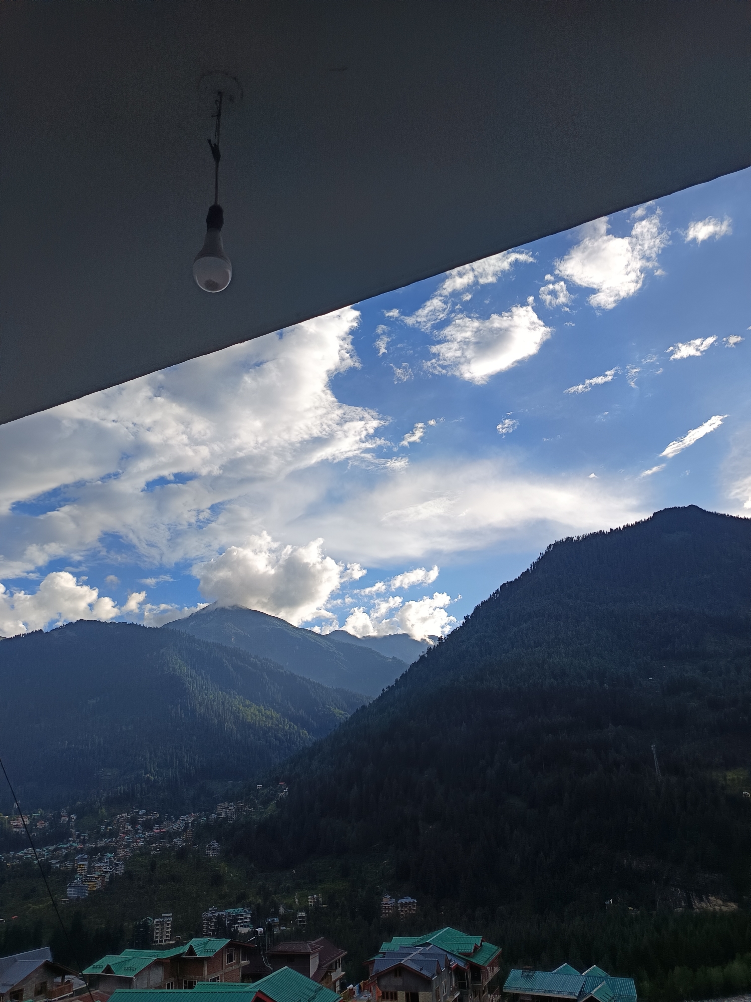 The image captures a scenic view of the mountains surrounding the town of Vashisht, India, with a portion of a building's roof and an overhead light fixture visible in the foreground. The sky is a vibrant blue, dotted with fluffy white clouds, some of which are catching the sunlight. The mountains are covered in a dense forest of dark green trees, with patches of buildings forming a small town nestled in the valley between the peaks. The architecture of the visible buildings suggests a rustic charm, with many featuring distinctively sloped, multi-colored roofs. The overall impression is one of natural beauty and a peaceful mountain village.