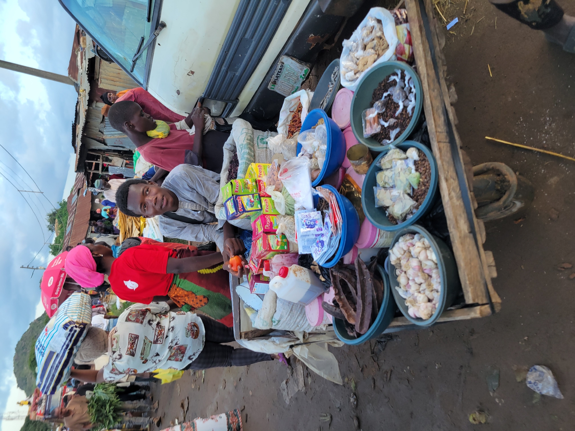 A young African male wearing a grey shirt and a yellow scarf around his neck is seated in front of a market stall in Gura Top, Nigeria. He is looking towards the right side of the frame, with his hands resting on the stall. The stall is laden with various food items, including bags of grains, dried beans, and what appear to be spices. In the background, other people are visible, as well as market stalls and makeshift structures. A white vehicle is parked to the right of the stall. The overall atmosphere suggests a busy marketplace.