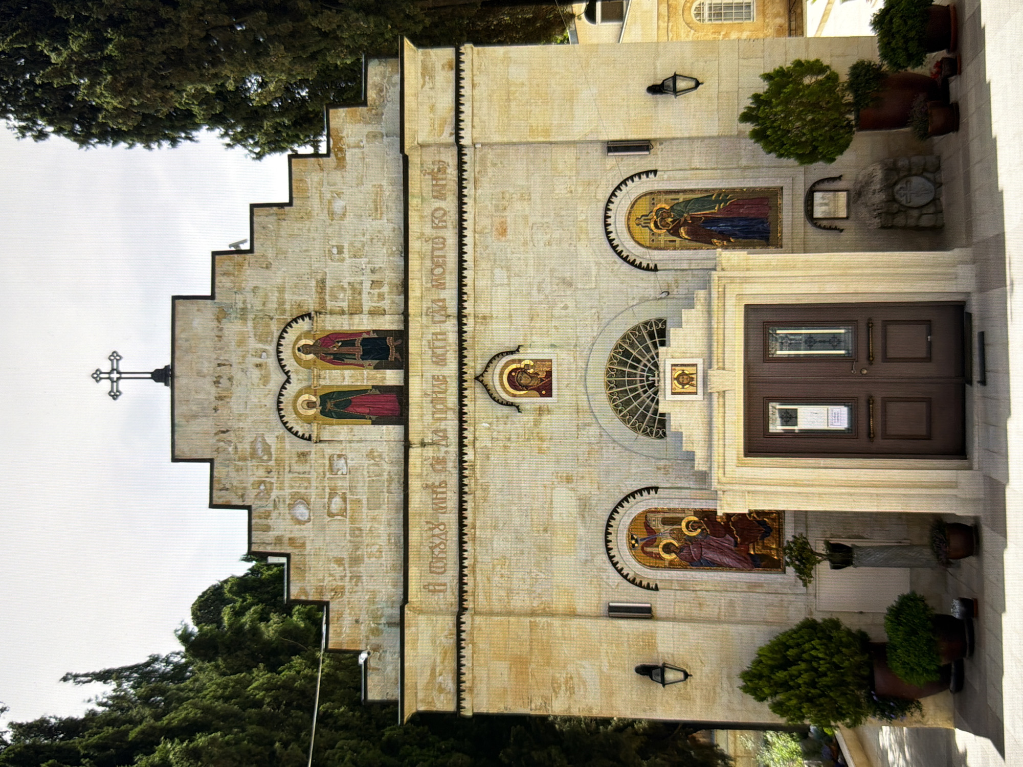 The image depicts a stone church with a stepped facade and a prominent cross on top. The facade is adorned with several mosaics featuring religious figures. There are two large doors at the entrance, flanked by potted plants. The church is surrounded by tall trees. The surrounding environment suggests it is an outdoor scene, possibly in a temperate climate. The image does not provide enough information to determine the specific location within Kyiv, Ukraine.