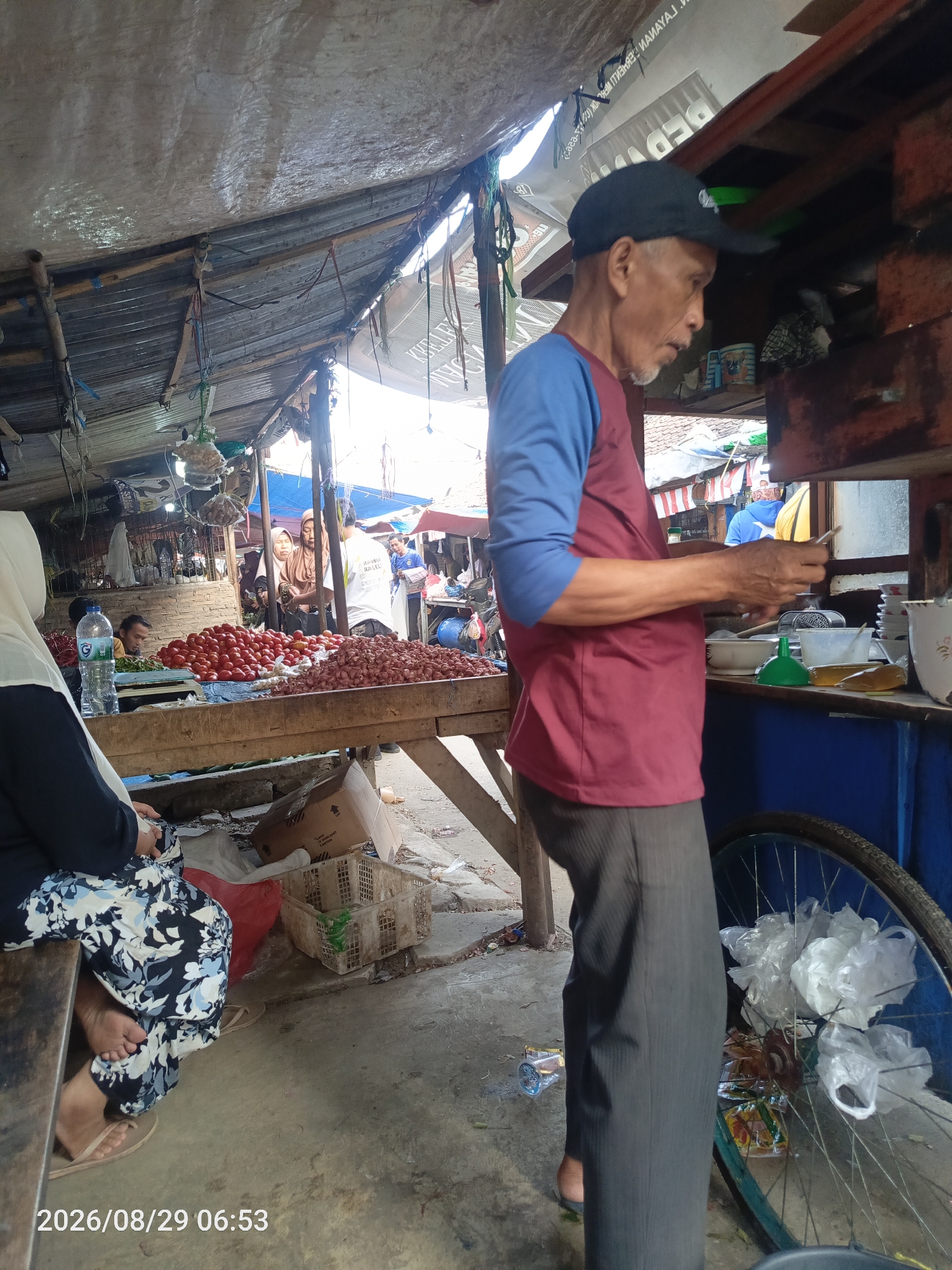 This image depicts a market scene in Sangiang, Indonesia, on August 29, 2026, at 06:53. In the foreground, an elderly man wearing a black baseball cap and a maroon and blue long-sleeved shirt stands near a food stall. He appears to be engaged in an activity at the stall, holding a small object in his hands. The stall itself is rustic, with a wooden counter and shelves displaying bowls, a green funnel, and a bottle of liquid. Behind him, a bicycle wheel with plastic bags tied to it is visible.
In the mid-ground, a woman wearing a white hijab and a patterned black and white dress sits on a wooden bench, facing away from the camera. To her left, a table is laden with fresh produce, including a large pile of red tomatoes and a mound of red onions. Various plastic baskets and a cardboard box are scattered on the ground. Further back, other market-goers and stalls are visible, suggesting a bustling environment, with some stalls covered by tarps or awnings. The overall environment appears to be an open-air market with a mixture of permanent and temporary structures.