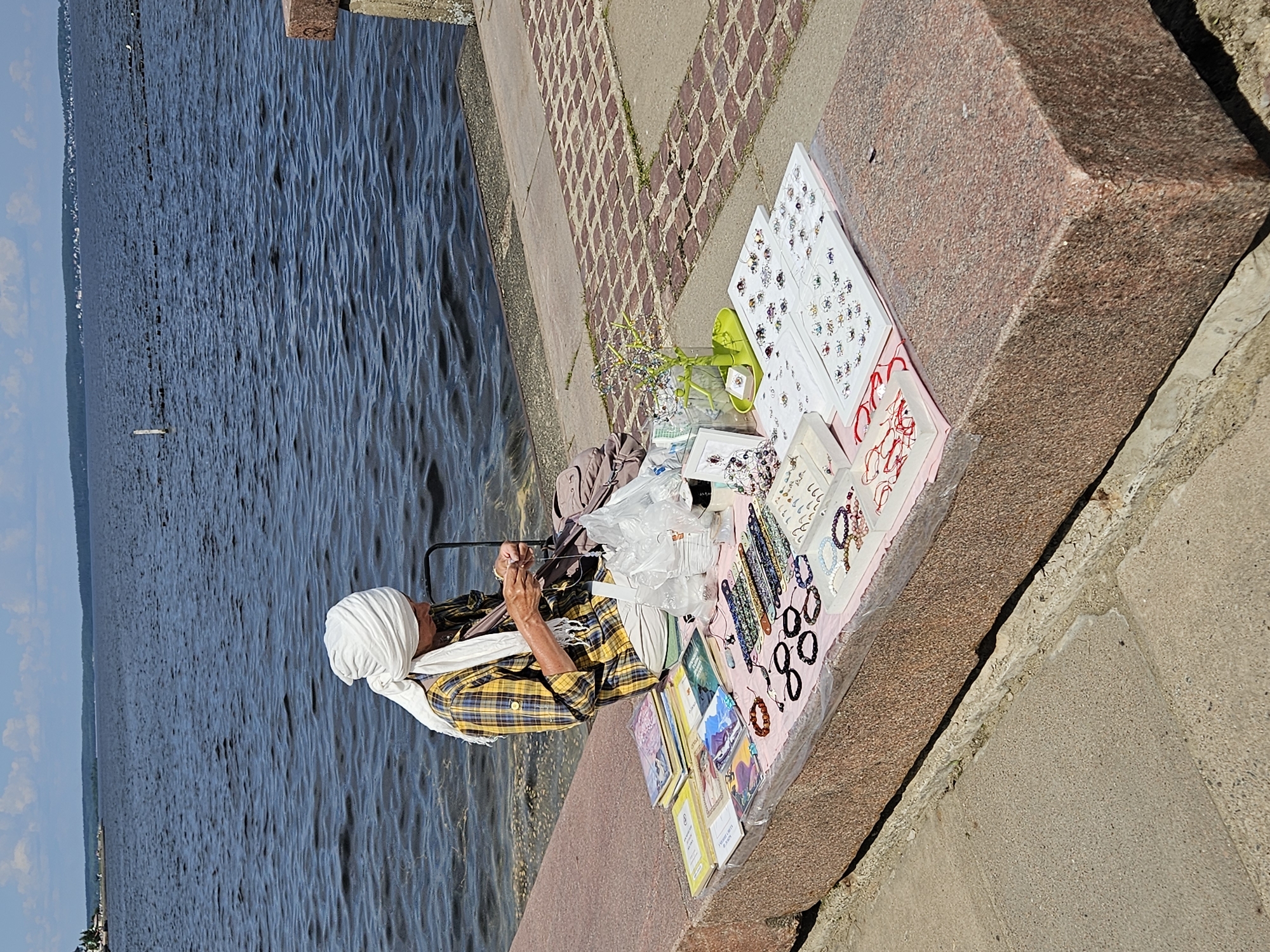 An outdoor, eye-level, medium shot captures a person sitting at a stone bench by the water's edge, creating jewelry. The person, wearing a white headscarf and a yellow and blue plaid shirt over light-colored pants, is actively engaged in stringing beads. Spread out on the bench before them is a collection of handmade jewelry, including bracelets and necklaces, neatly arranged on pink and white displays, along with some books and plastic bags. The background consists of a vast expanse of dark, rippling water under a partly cloudy blue sky, suggesting a warm, sunny day. The foreground shows a textured stone bench and a paved walkway.