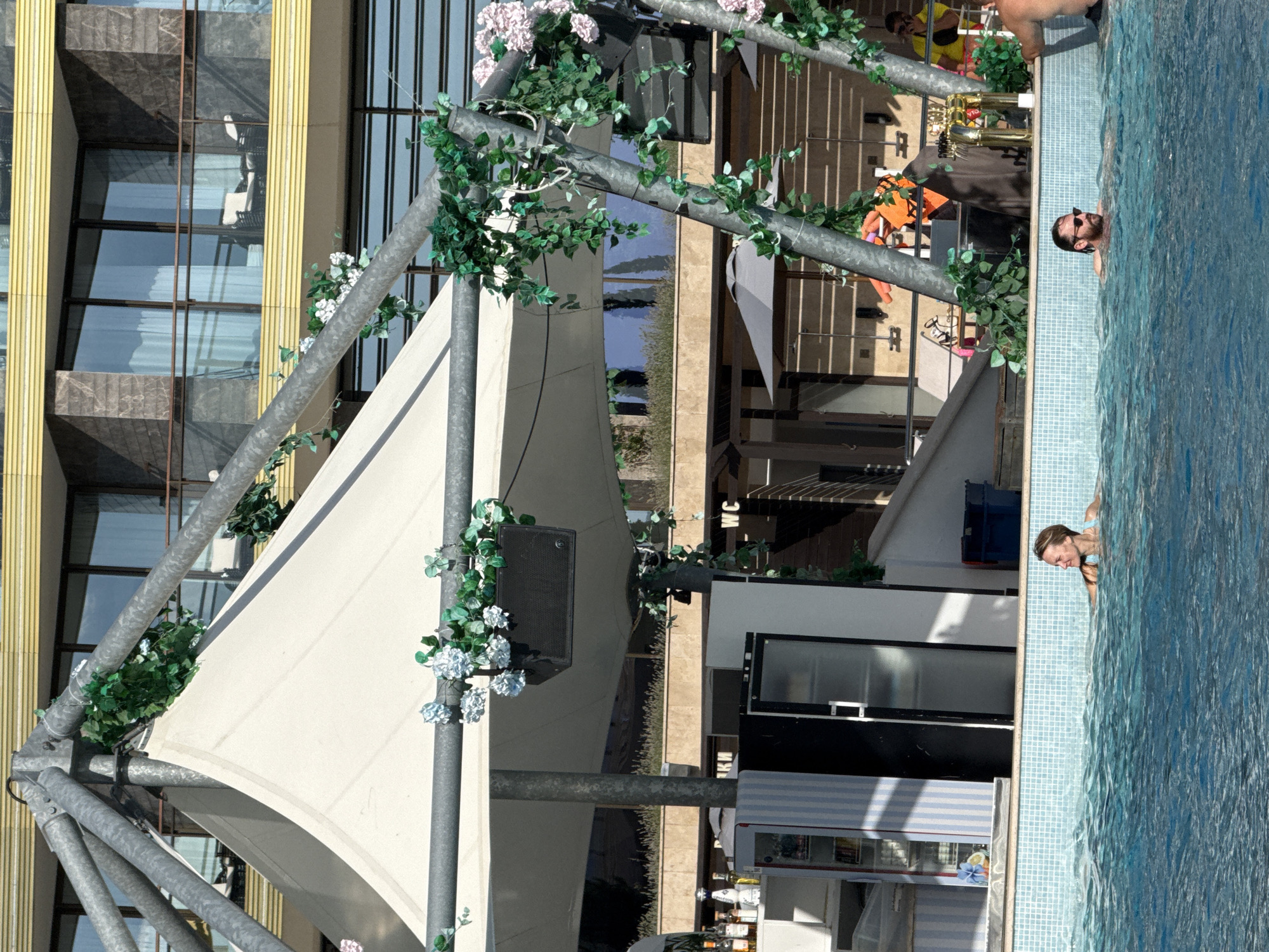 In the city of Simeiz, Ukraine, a refreshing pool scene unfolds under a bright sky, with a modern building forming a backdrop. At the center, a white tent structure, supported by metal poles, offers shade. Greenery and artificial flowers adorn the poles, adding a touch of nature. To the left, a bar area features shelves stocked with bottles and refrigerators. Two individuals are visible in the pool: a woman with blonde hair, wearing a blue swimsuit, is swimming, while a man with a beard and sunglasses, also in swimwear, floats nearby. The pool itself is tiled with blue and white squares, creating a vibrant mosaic. In the background, a multi-story building with balconies and large windows stands prominently, its architecture reflecting the contemporary style of the resort. The scene captures a moment of relaxation and enjoyment at a poolside establishment.