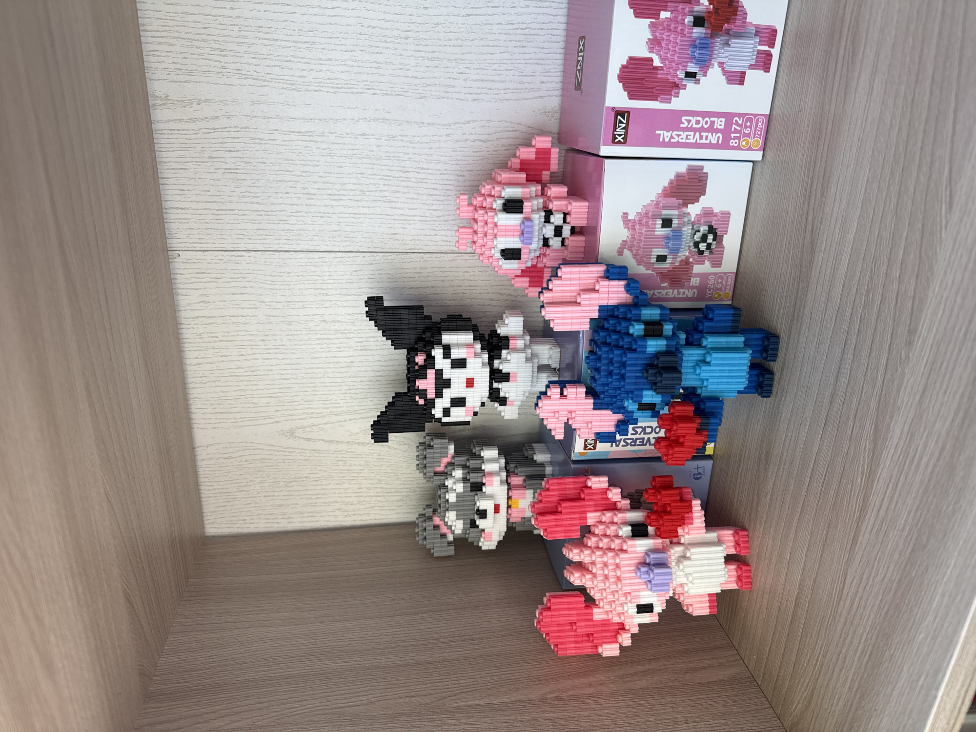 The image shows several assembled XINZ Universal Blocks toys of cartoon characters and their respective boxes, stacked and positioned inside what appears to be a wooden shelf. There are four assembled figures, two in shades of pink, one primarily blue, and one featuring black and gray. The boxes are pink and white, stacked behind the toys. The scene is lit well, and the background features a wooden texture, indicative of the shelf's material.