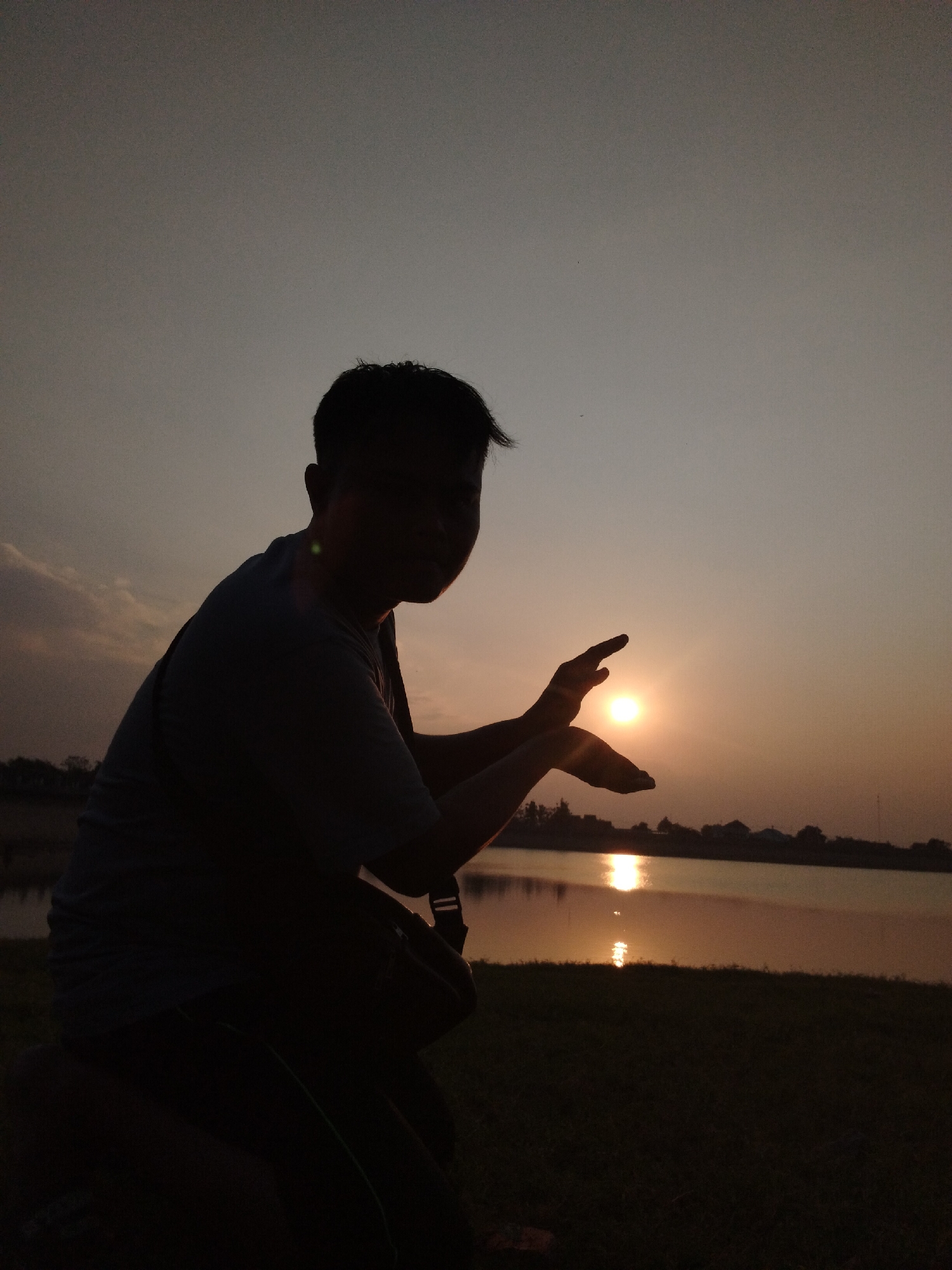 This is a silhouette photograph taken at sunset in Sambirejo, Indonesia, likely near a body of water. A person is positioned in the foreground, sitting on an unseen surface and facing towards the right of the frame. The individual is silhouetted against the warm, hazy sky, with the sun prominently visible in the middle ground. The person's hands are raised, with their fingers positioned in a way that appears to be framing or interacting with the setting sun. One hand is cupped, and the other is held open, with the sun appearing to be partially enclosed within the space between them.
In the background, a calm body of water reflects the colors of the sunset, creating a shimmering path of light on its surface. The far bank of the water features a silhouetted treeline and what appear to be buildings, indicating a populated area nearby. The sky itself is a gradient of pale gray at the top, transitioning to warmer orange and yellow hues near the horizon where the sun is located. The overall impression is one of a serene and contemplative moment captured during twilight.