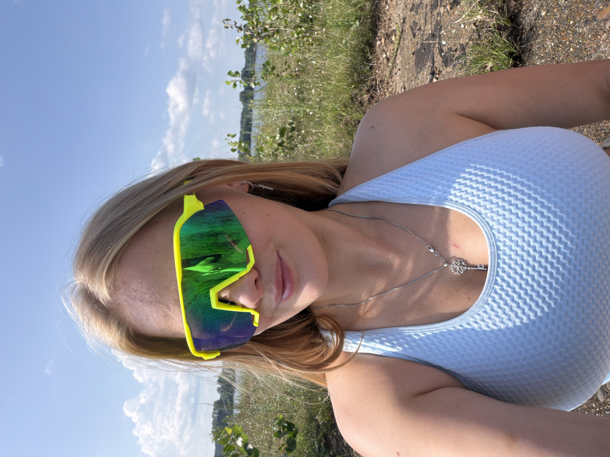 In a medium shot, a woman with blonde hair, wearing bright yellow-green mirrored sunglasses and a light blue textured tank top, is pictured outdoors with a lake and green foliage in the background. Her reflection is visible in the sunglasses, showing a green and blue pattern. She is wearing a silver necklace with a key-shaped pendant. The scene appears to be a sunny day with scattered clouds in a blue sky, and the ground is covered in dirt and small rocks. There is no specific identifiable landmark or feature within the image that allows for definitive identification of the location within Chaikovsky, Russia.
