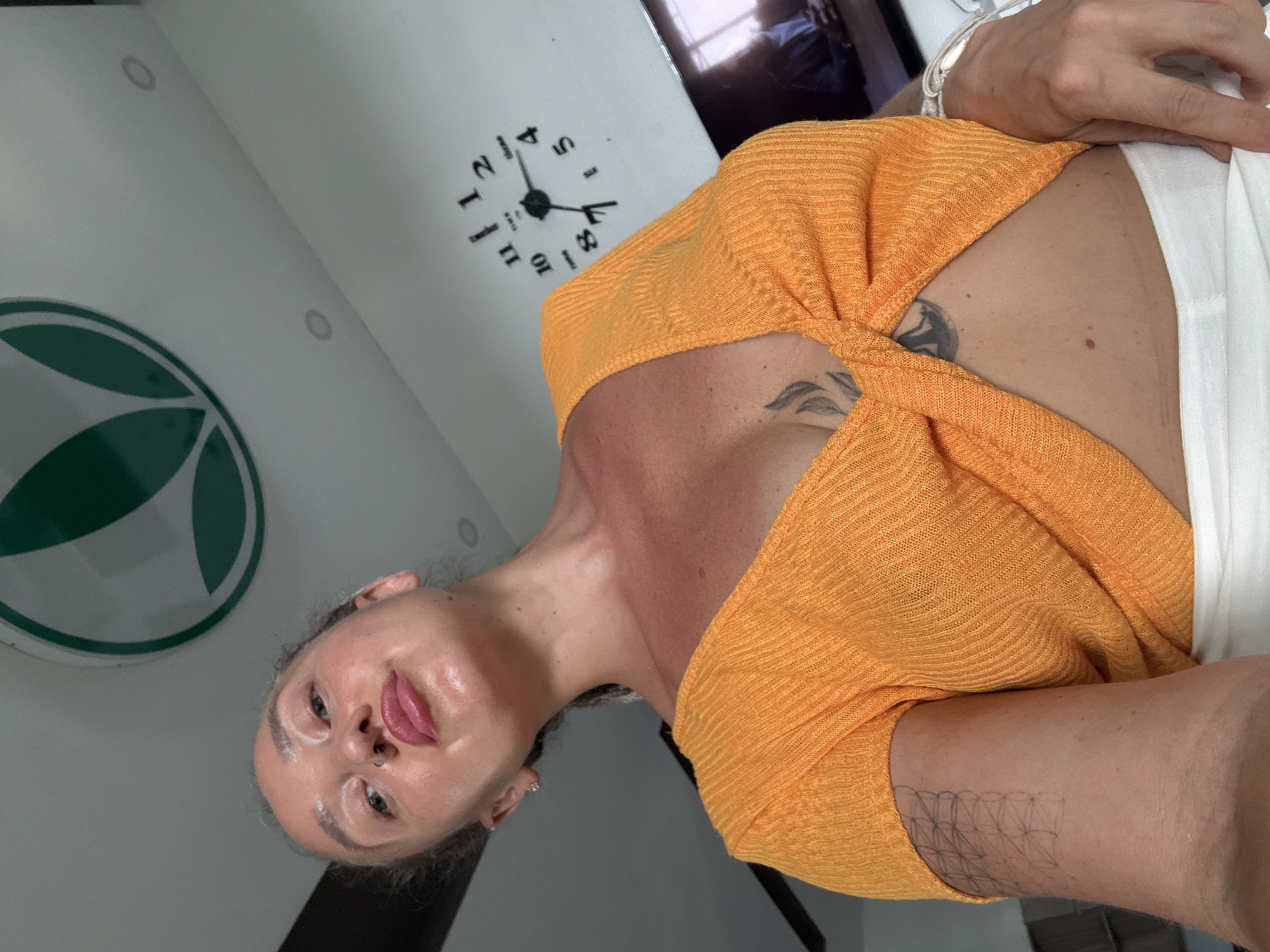 A young woman is shown from the chest up, with her head tilted back. She is wearing an orange, twisted top, and her hair is pulled back. There is a tattoo on her chest and arm. A clock is visible on the wall behind her, as well as a circular logo with green leaves. The lighting suggests it is daytime. The image appears to be a selfie. There is no information in the image to suggest this is located in Bac Nha Trang Ward, Vietnam.
