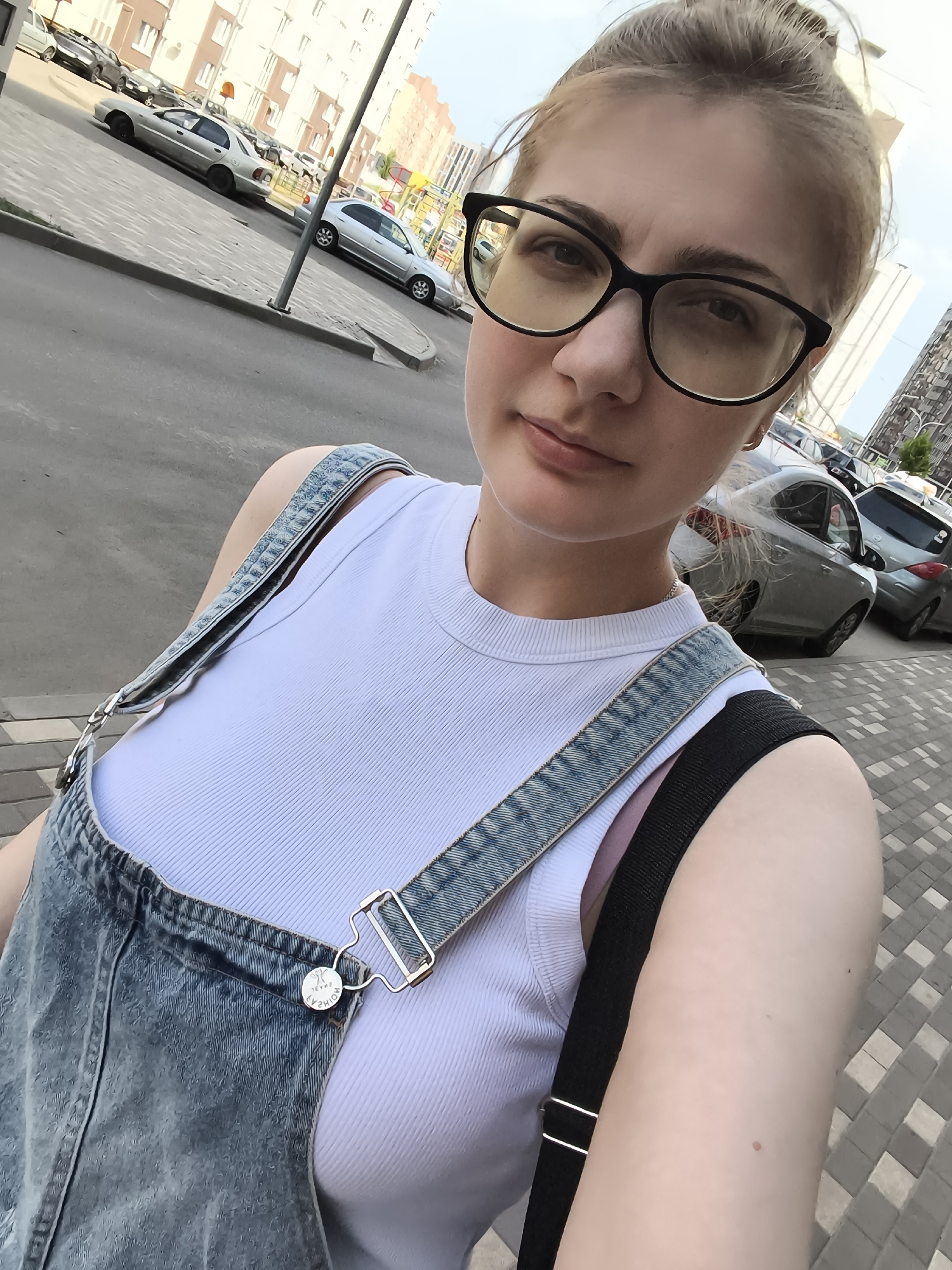 A young Caucasian woman with blonde hair pulled into a bun and wearing black-rimmed glasses is taking a selfie outdoors. She is dressed in a white ribbed tank top and denim overalls, with a black strap of a bag visible across her shoulder. The background shows a street with parked cars and a sidewalk with a patterned paving. Buildings are visible in the distance. The lighting suggests it is daytime.