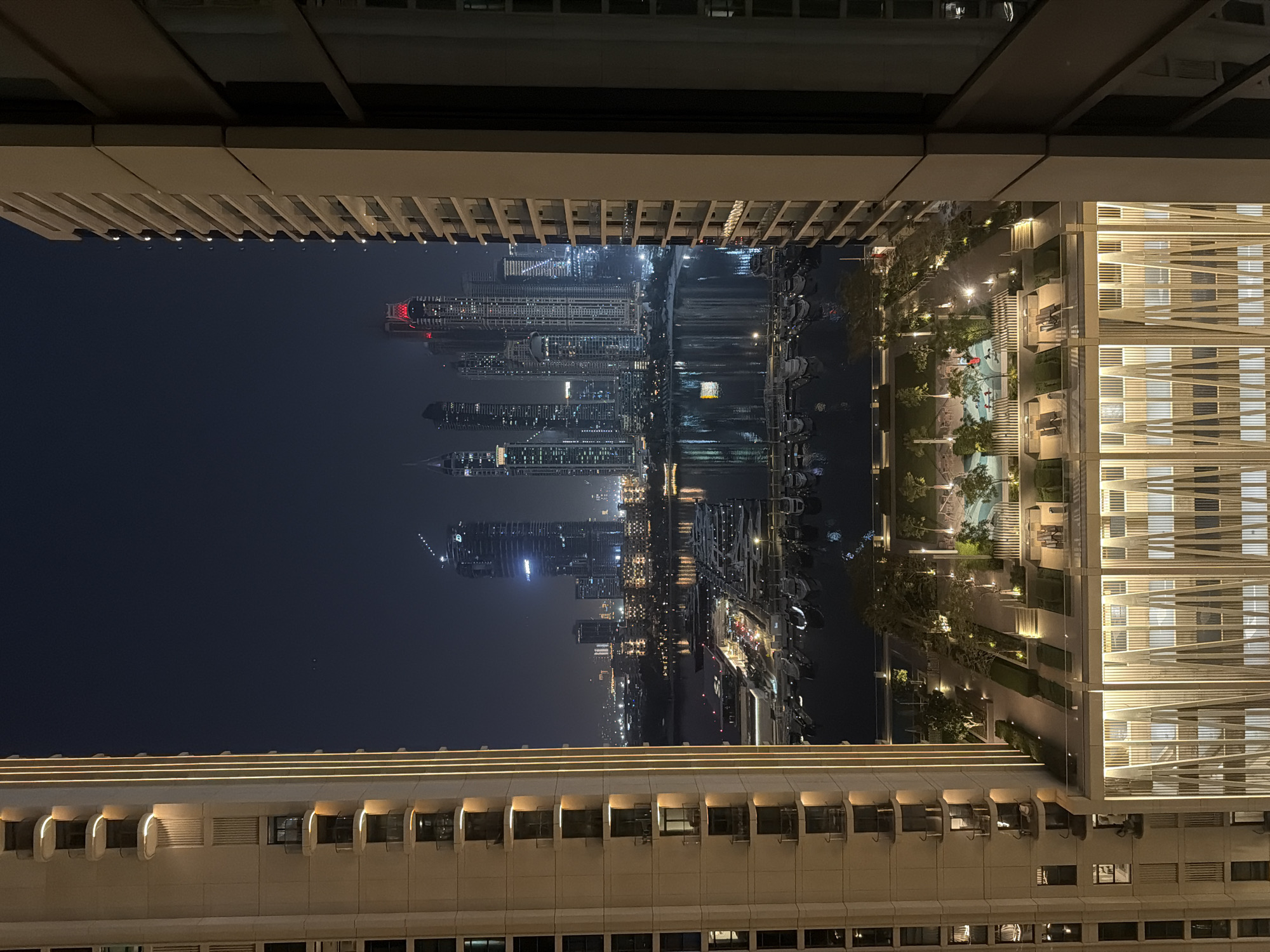 This is a nighttime exterior, medium shot photograph taken from a high vantage point overlooking a waterfront development in Dubai, United Arab Emirates. The foreground is dominated by the sides of two modern high-rise buildings. The left building is a beige-colored structure with balconies and vertical accent lighting. The right building is a darker colored structure, also with balconies.
Between these buildings, the scene opens up to a view of a cityscape and a marina. In the midground, there is a body of water, likely a canal or bay, filled with numerous docked yachts and boats. The water reflects the lights from the surrounding buildings and boats. A promenade or walkway runs along the water's edge, with some lights visible.
Further in the distance, the iconic Dubai skyline is visible, characterized by numerous skyscrapers of varying heights and architectural designs. Several buildings are illuminated with colored lights, particularly red on the tops of some towers. The sky is dark, indicating night.
In the lower part of the frame, between the two foreground buildings, there is a landscaped area with trees, pathways, and seating areas. There is also a swimming pool visible in this area. The architectural style of the buildings suggests a luxurious, contemporary urban environment, consistent with prominent areas in Dubai such as Dubai Marina or Business Bay.