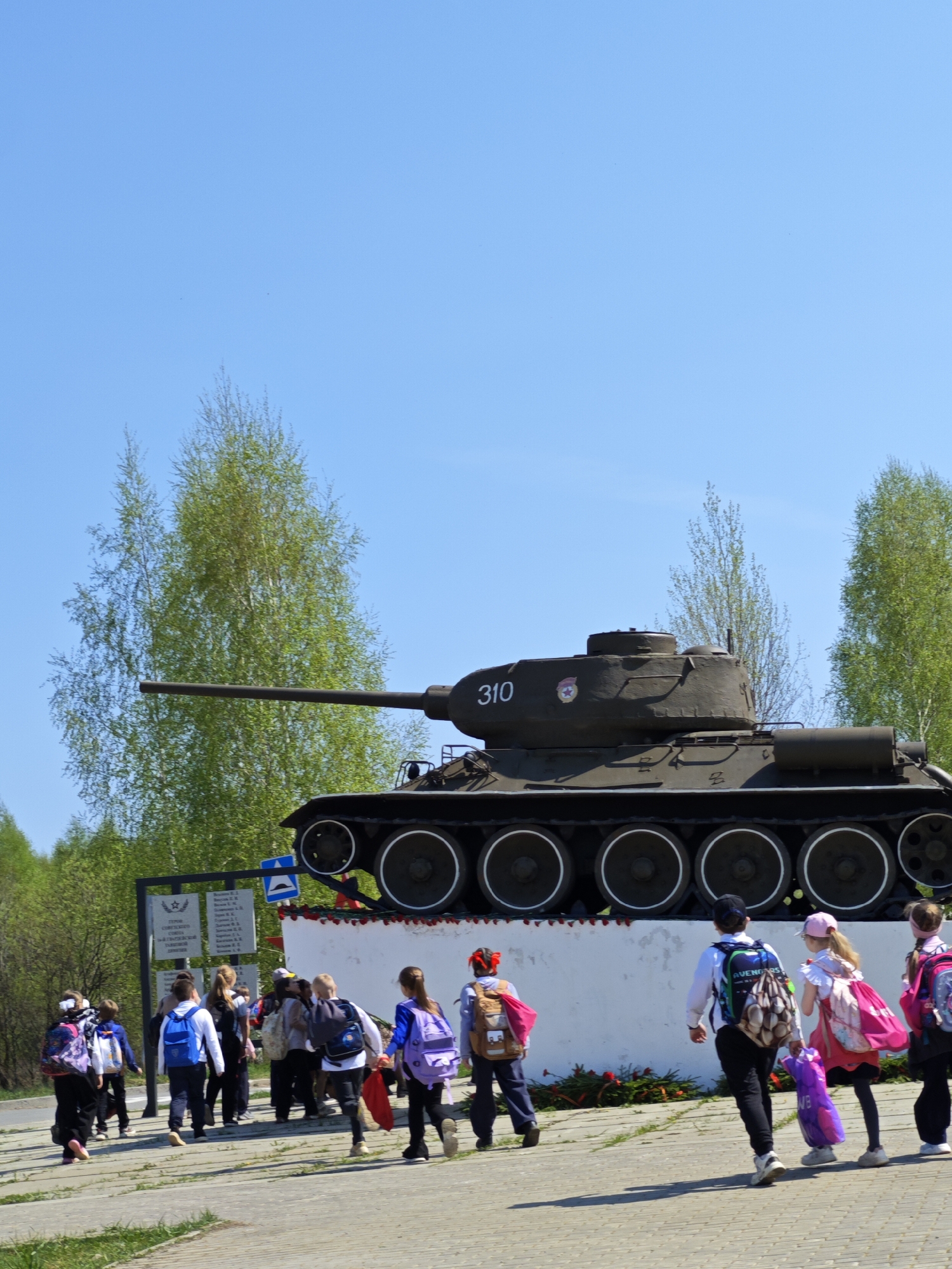 A group of children with backpacks walk past a Soviet T-34 tank memorial in the settlement of Марковский, Russia. The tank, marked "310", is mounted on a white platform adorned with red flowers. A sign is visible behind the group of children on the left of the picture. The sky is bright and blue. Trees are visible in the background. The ground has a concrete sidewalk.