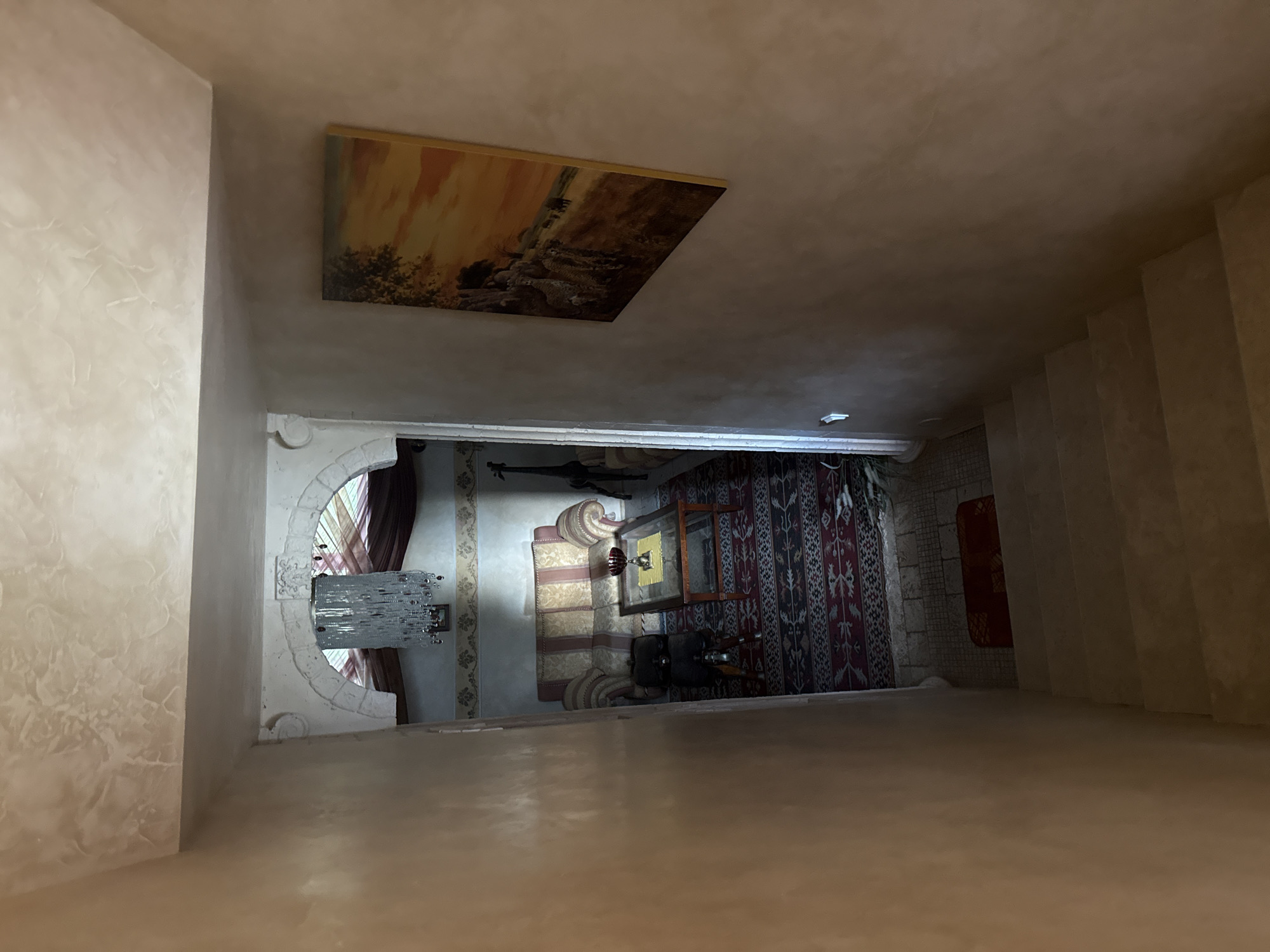 The image shows a view from the top of a staircase looking down into a room. The staircase is made of concrete and is illuminated by natural light. The room below is decorated in a traditional style, with a patterned rug, a sofa, and a coffee table. Above the sofa, there is a chandelier. A painting of animals in a savanna landscape hangs on the wall to the right of the staircase. The walls of the staircase and the room are painted in a light, earthy tone. The overall impression is one of an old, well-decorated house. The city of Yukki, Russia, is not visible in the image.