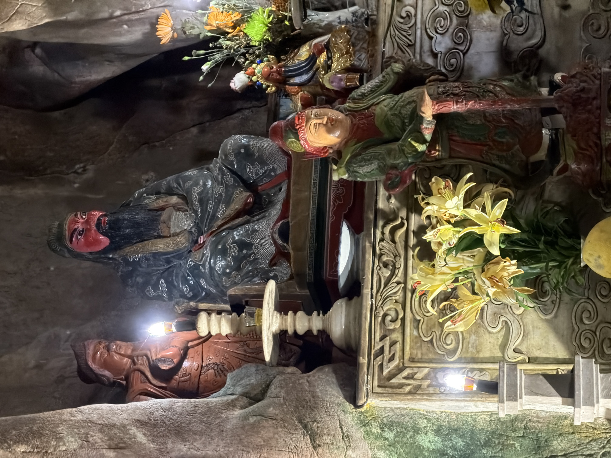 The image shows a shrine within a cave or grotto in Đà Nẵng, Vietnam. There are multiple carved figures. One figure is large with a black beard and red face. Another figure is lying prone in a green outfit. There are also yellow lilies in a vase. The shrine is ornately decorated and set on a stony surface. Artificial lighting is present. The surrounding area appears to be cave walls.