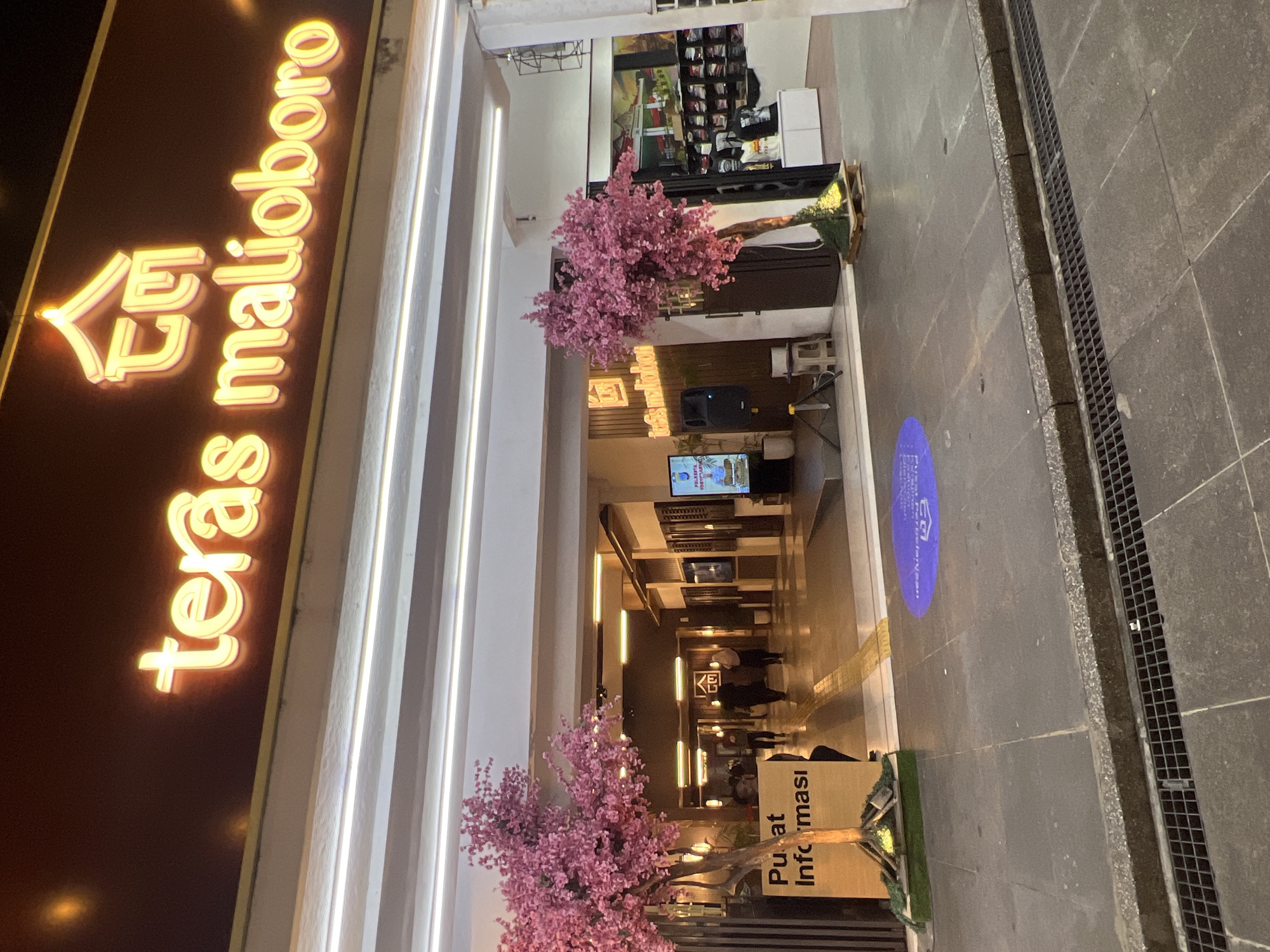 The image depicts the entrance to "Teras Malioboro," a commercial establishment in Yogyakarta, Indonesia. The facade of the building features a prominent neon sign with the text "teras malioboro" in illuminated yellow letters, accompanied by a stylized architectural logo. In front of the entrance, two artificial trees with pink blossoms are placed, one on the left and one on the right.
To the left, a sign reads "Pusat Informasi," indicating an information center. Several people are walking through the arched entryway into the building. The interior appears to be a marketplace or mall with shops and displays. On the right side of the entrance, a screen displays information about "POLRES YOGYAKARTA". A blue circular social distancing marker is visible on the ground in front of the entrance. The ground is paved with dark tiles. A grate covering a drainage channel runs along the bottom of the image. The scene is illuminated by artificial lighting, suggesting it is nighttime or an indoor environment.