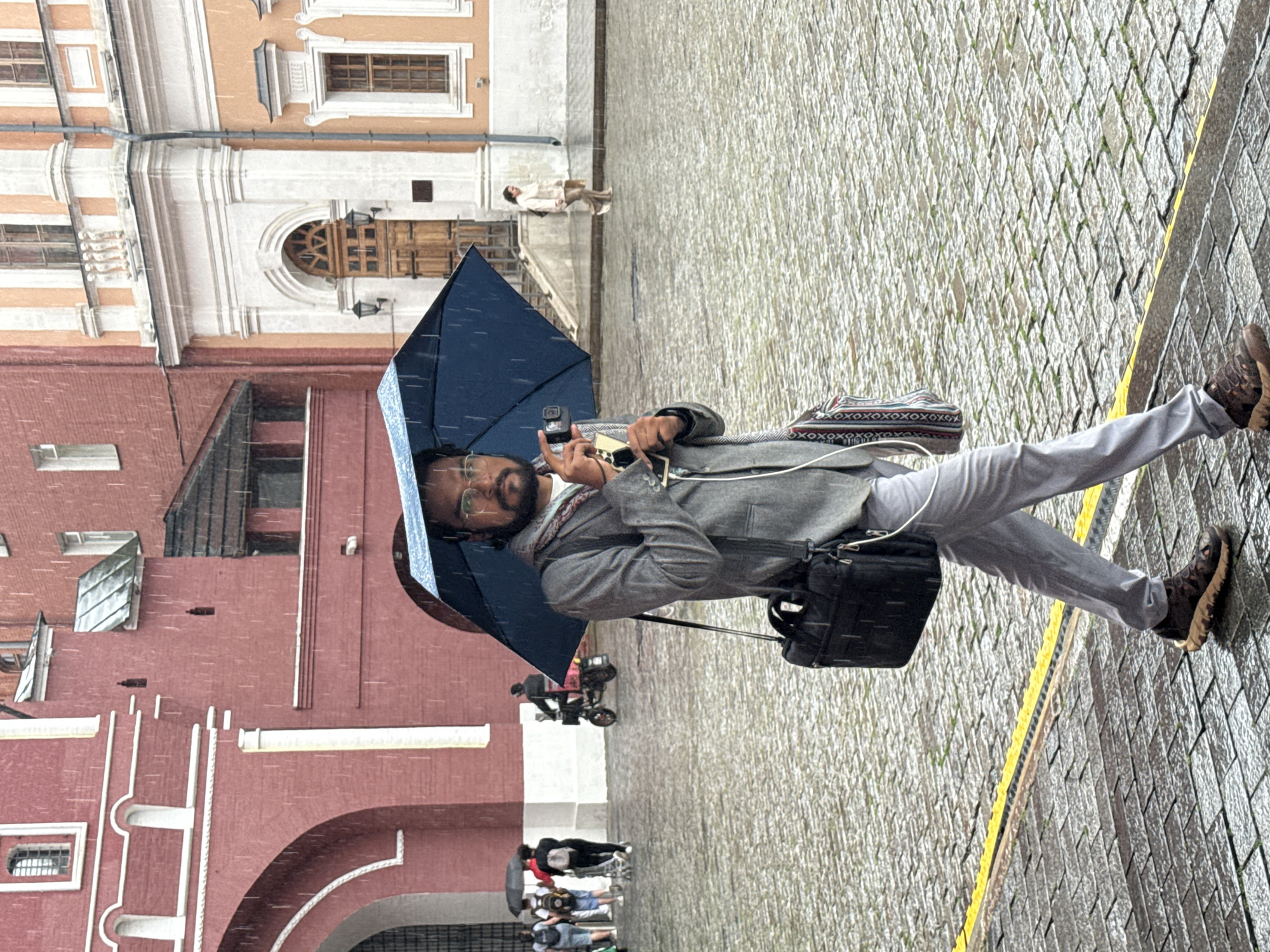 A man walks under a blue umbrella in the rain on a cobblestone street in Moscow, Russia. He is wearing glasses and a gray jacket, and he is carrying a black briefcase. In the background, other people are walking and a person is riding a scooter. The buildings in the background are historic and appear to be made of red brick and white stone. The street is wet and reflects the light.