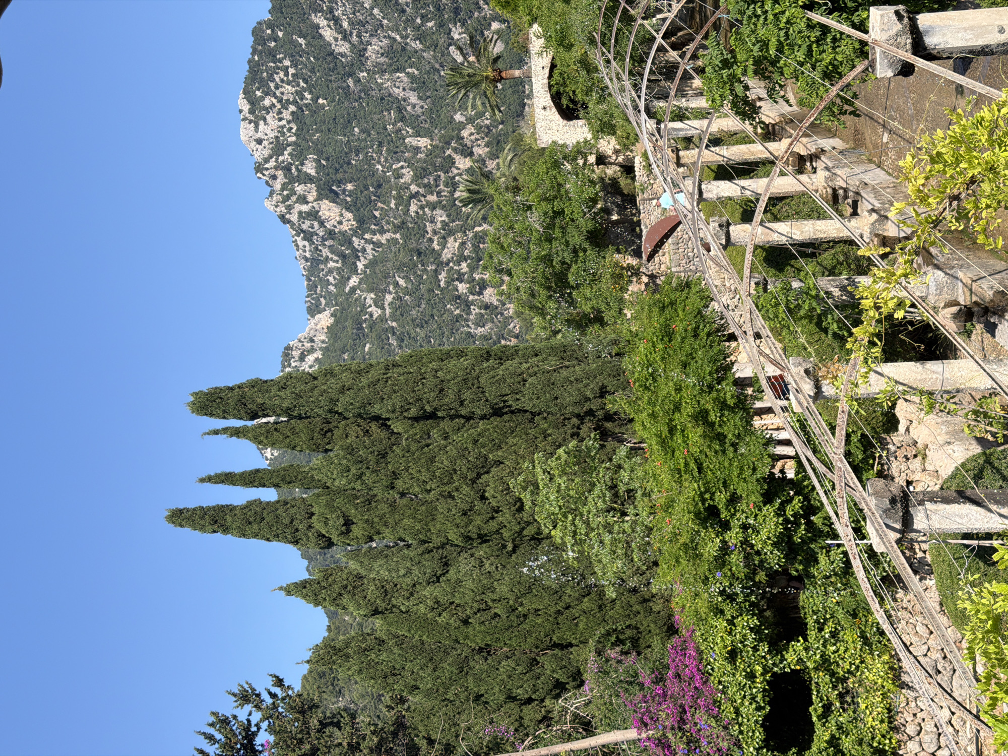 The image shows a view of a stone structure overgrown with greenery and flowers, possibly located in or near the city of Bunyola, Spain, given the mention of its location. A mountainside looms in the background under a clear, blue sky. A series of tall, thin, evergreen trees stand vertically in the midground. In the foreground, there is an arched, metal structure over a series of stone steps and vegetation. The scene is illuminated by bright sunlight, with no people visible.