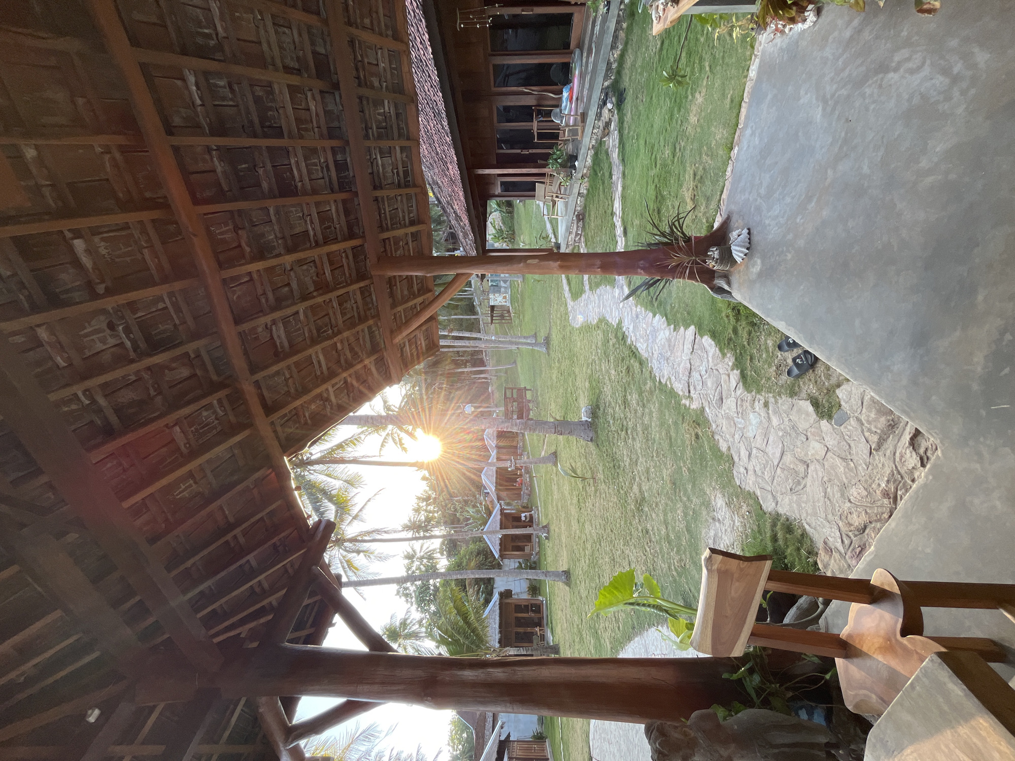 The image captures a tranquil outdoor scene at a resort in Legonipah, Indonesia, during what appears to be sunset. The foreground is dominated by a wooden roofed structure, supported by thick wooden pillars, offering a view of a well-maintained lawn dotted with coconut palm trees. A pathway of irregular stone slabs meanders through the grass, leading towards several small wooden bungalows with thatched roofs, visible in the mid-ground. The sun is setting behind the palm trees, casting a warm, golden glow and creating sunbursts through the foliage. A wooden chair is positioned in the foreground, facing the lawn and bungalows. The overall atmosphere is serene and tropical.