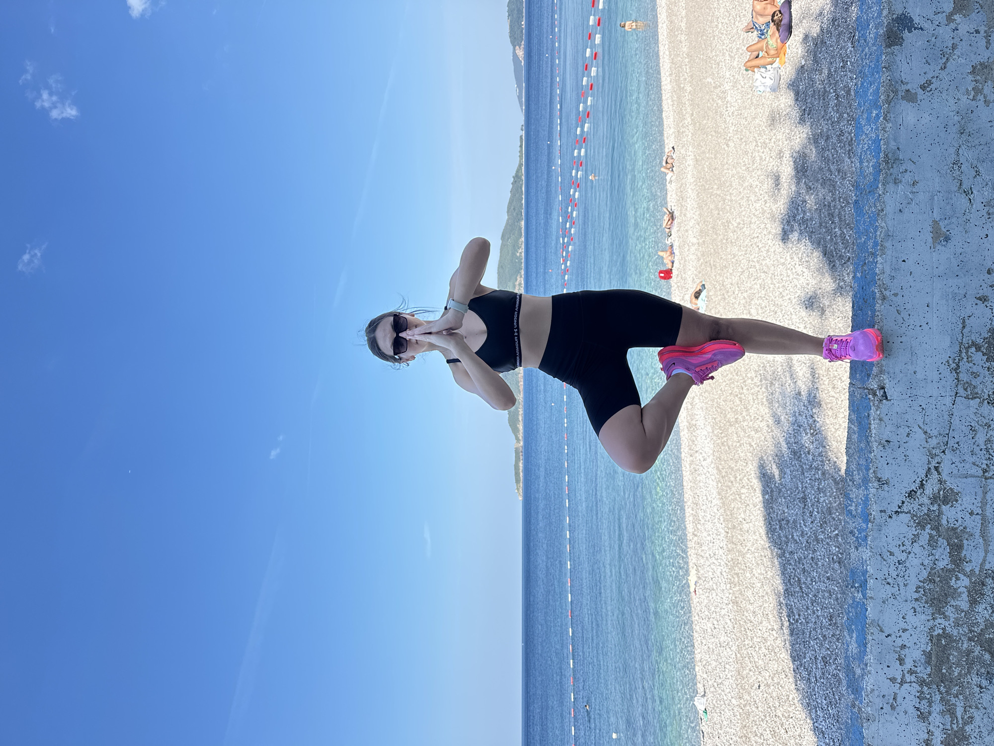 A young woman is captured in a tree pose on a concrete wall overlooking a beach in Bečići, Montenegro. She is wearing a black sports bra and black bike shorts, with bright pink athletic shoes. Her hands are pressed together in front of her chest, and she is looking forward with sunglasses on. The beach behind her is covered in white pebbles, with a clear blue Adriatic Sea stretching to the horizon. In the distance, a hilly coastline is visible. Several people are sunbathing on the beach, and a few are in the water. Red and white buoys mark off an area in the sea. The sky is a clear, bright blue with a few wispy clouds. The sunlight casts shadows on the wall and the beach.