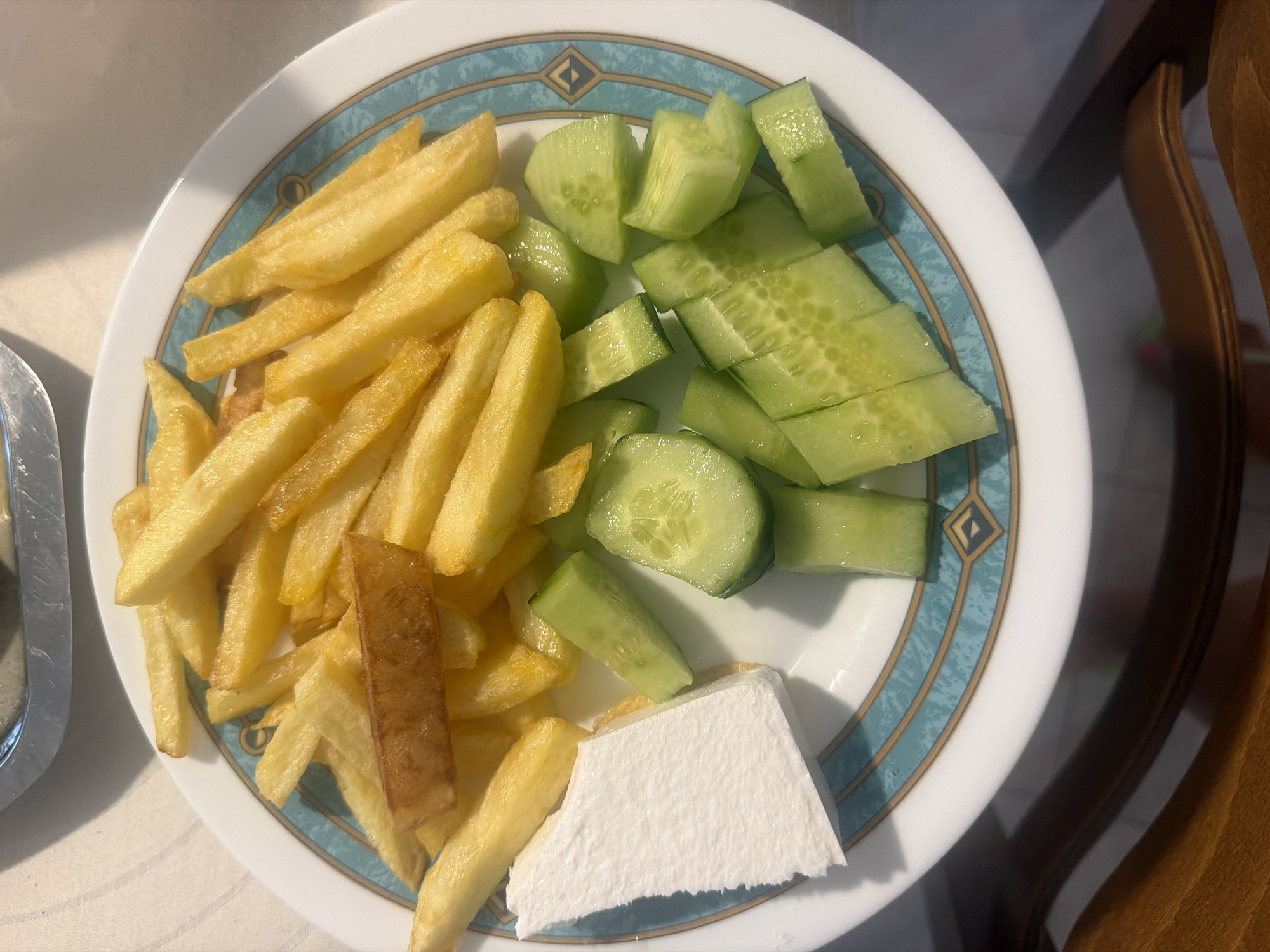 On a white plate with a turquoise and gold rim, a meal is served. The plate contains a pile of golden-brown french fries, a rectangular piece of white cheese, and sliced cucumbers. The cucumbers are cut into bite-sized pieces and appear fresh and slightly glistening. The scene is captured from a top-down perspective, with the plate resting on a light-colored surface. The background is blurred, but suggests an indoor setting with a glimpse of what appears to be a metal serving dish at the top. The overall impression is of a simple, fresh meal. This image was taken in Mandra, Greece.