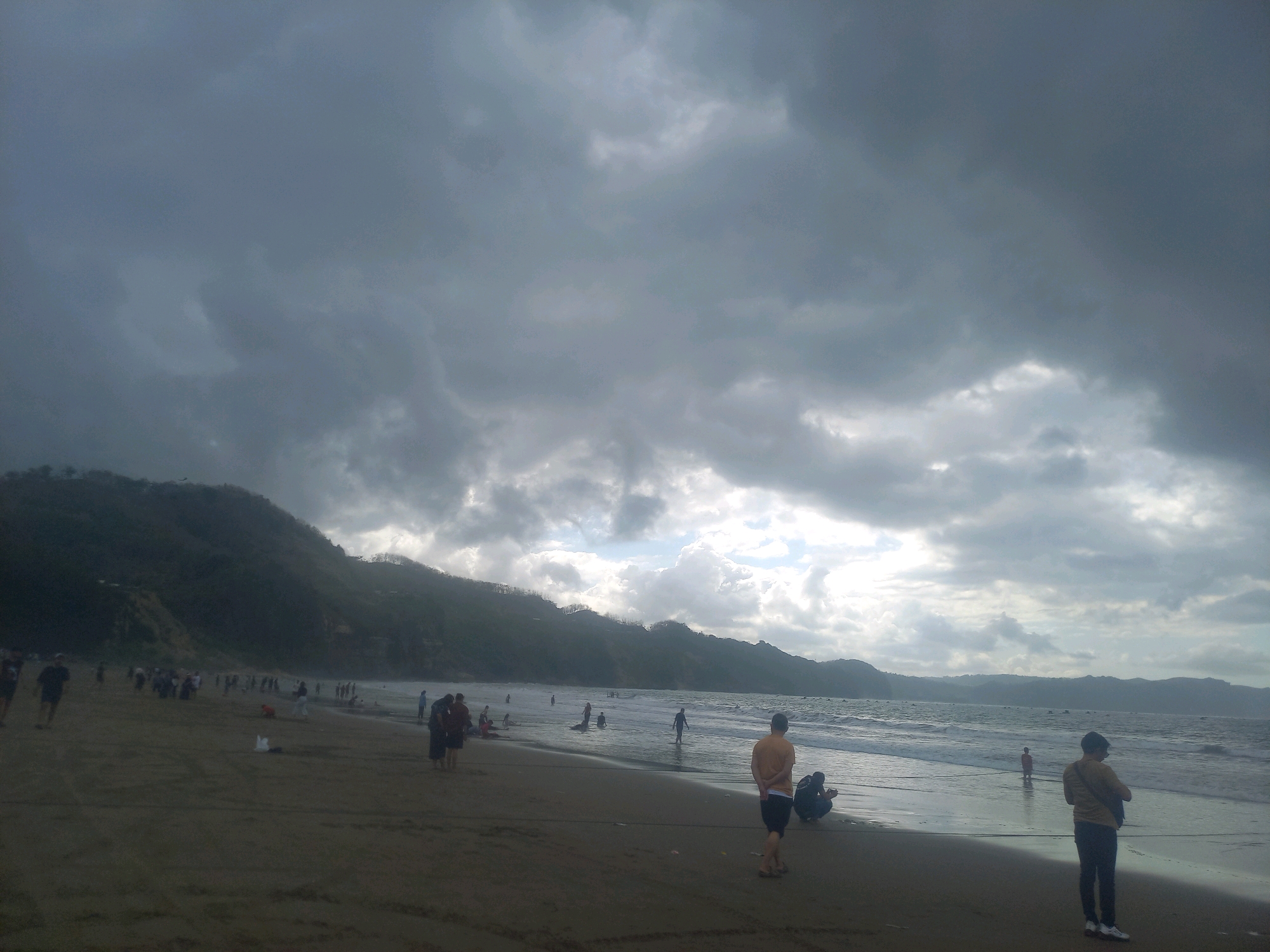 The image depicts a crowded beach scene with a dramatic, cloudy sky. Hills and mountains line the horizon behind the ocean, which features moderate waves. Numerous people are scattered across the sandy beach, some walking, others standing or sitting. A few individuals are visible in the shallow water. The overall impression is one of a busy recreational area under overcast conditions, possibly before or after a rain shower. The location is identified as a beach in Unknown, Indonesia.