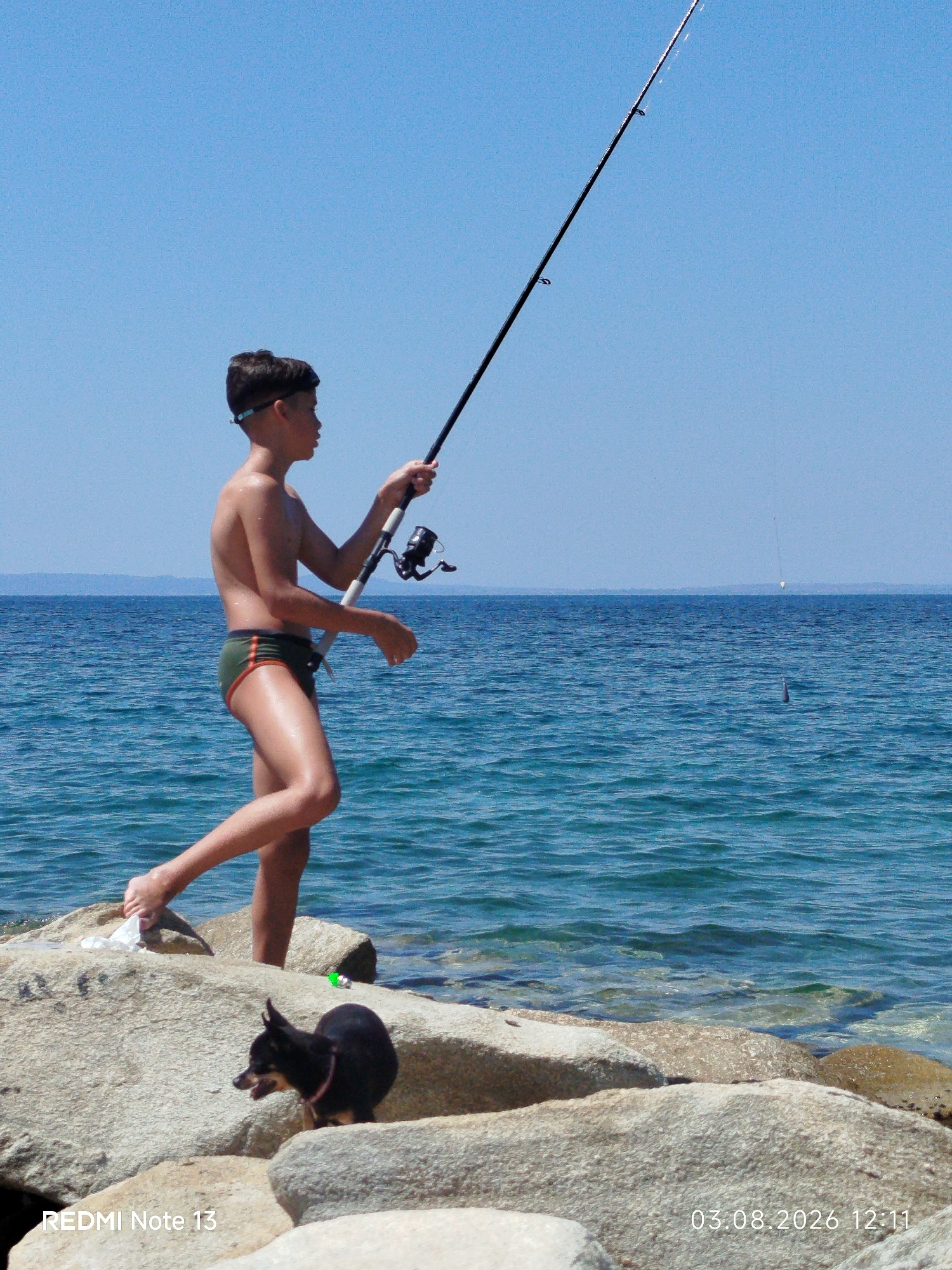 A young boy wearing swimming trunks and swimming goggles stands on rocks by the sea, holding a fishing rod and casting it into the water. A small black dog is also visible on the rocks in the foreground. The sea is blue and calm, with a clear sky above. The location appears to be a rocky coastline in Sithonia Municipal Unit, Greece, based on the timestamp and the distinctive scenery.
