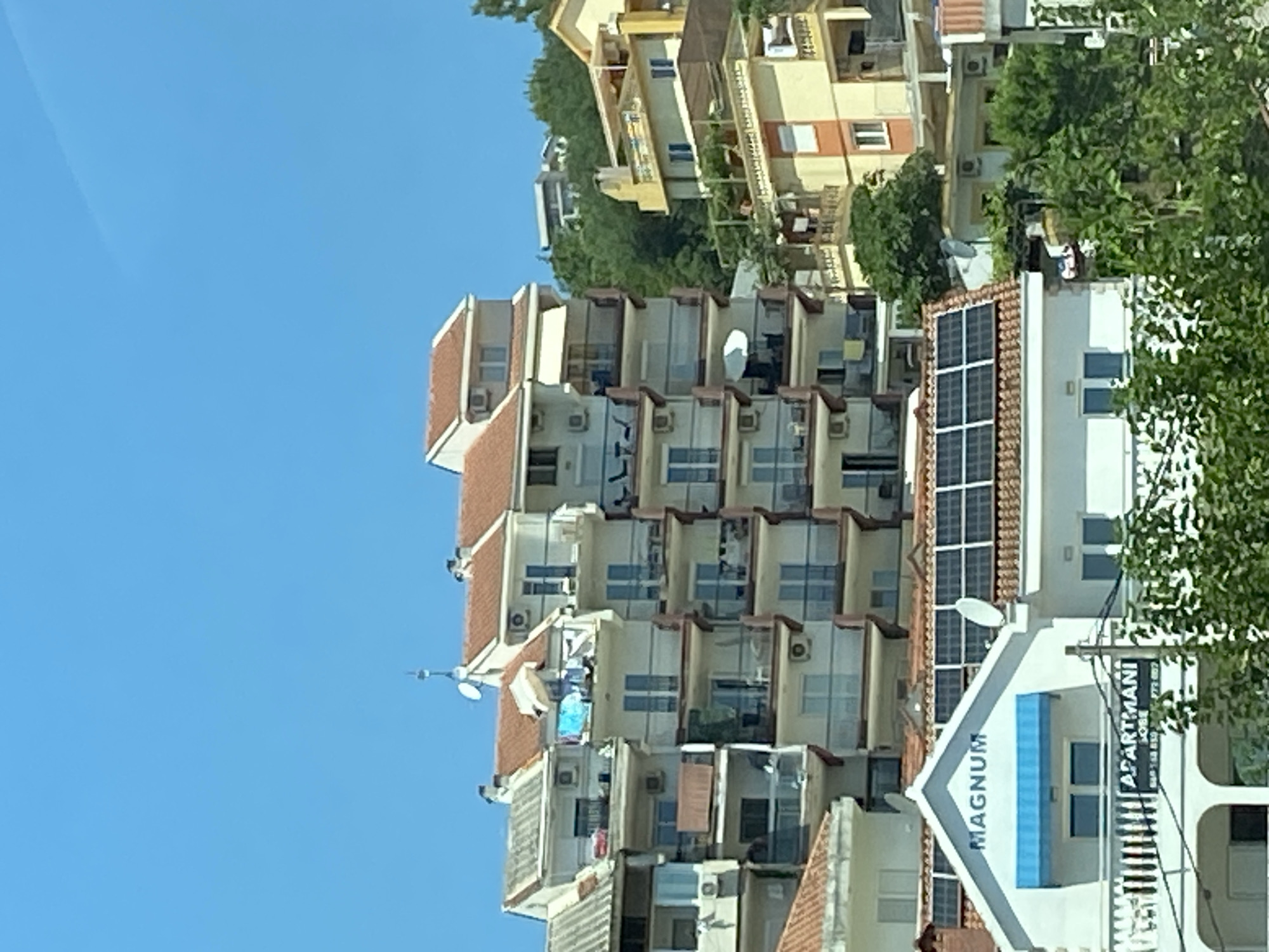 The image shows a multi-story apartment building with red tiled roofs in Meljine, Montenegro. The building is packed tightly with other similar structures, suggesting a dense urban environment. Several balconies are visible, some with chairs and tables, and laundry is hanging on one. Solar panels are installed on the roof of one section of the building. A sign on the front of this section reads "MAGNUM APARTMANI". The sky is clear blue, and lush green trees are visible in the foreground and background. No people are clearly visible in the image.