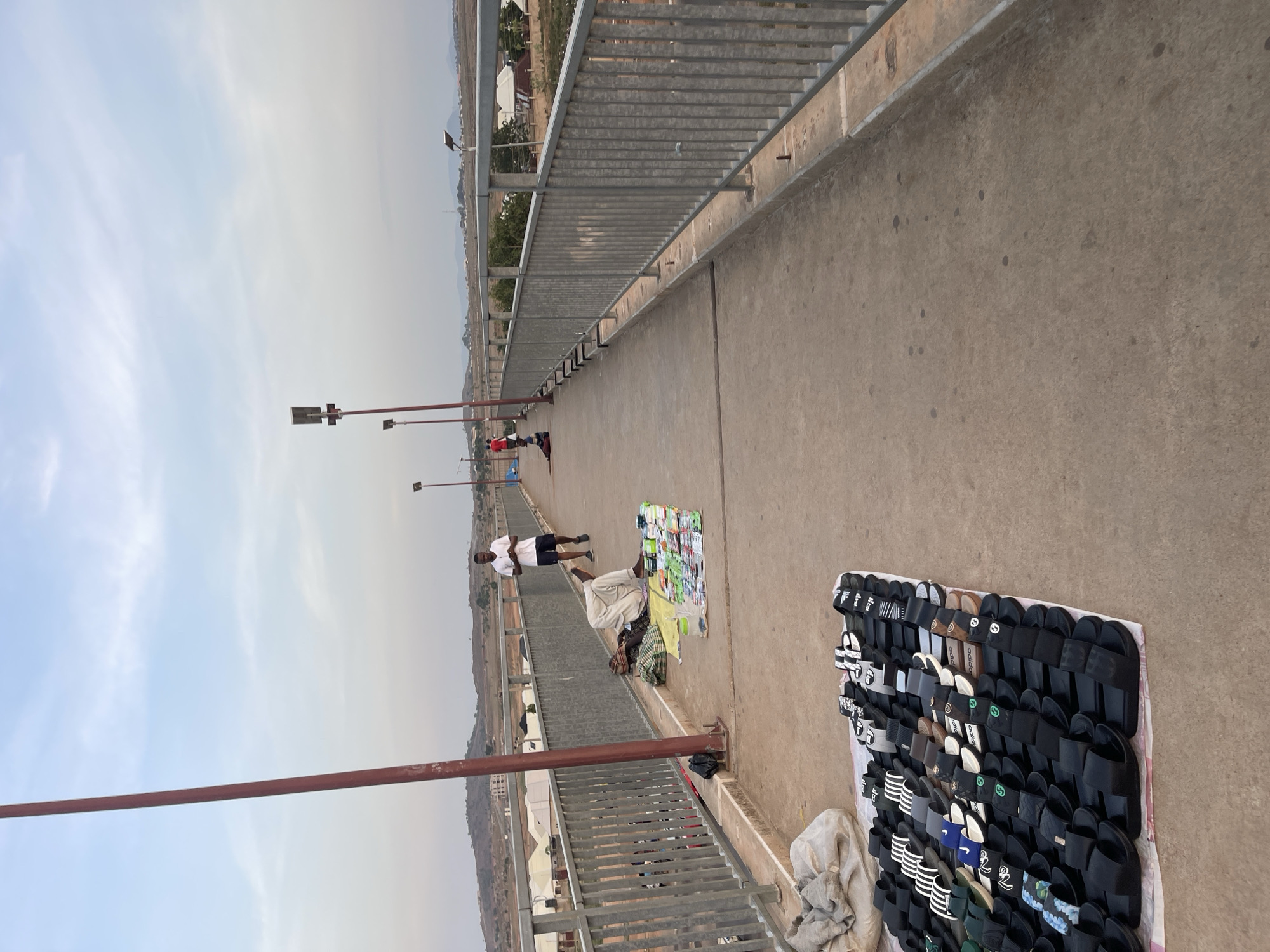 The image captures a scene on a bridge in Galadimawa Village, Nigeria. The bridge features metal railings and a concrete walking surface.
Several individuals are present. A man in a white shirt and black shorts stands near a collection of clothing and other items spread on a sheet. Another man stands further down the bridge.
Two displays of sandals are presented on the right. Streetlights are visible along the bridge. The surrounding environment is characterized by sparse vegetation and residential buildings in the distance. The sky is overcast.