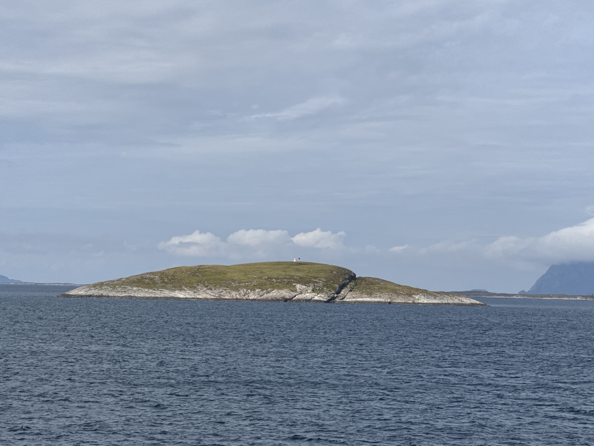 This is a medium-distance, eye-level, outdoor, horizontal, daytime shot of a small, green, rocky island in the sea. The island is covered with short, green grass and has a rugged, rocky shoreline. At the highest point of the island, there is a small, red and white structure that appears to be a lighthouse or navigational marker. A dark flag is flying next to it. The sea is a deep blue with moderate chop and ripples. The sky is overcast with scattered clouds and a pale blue hue. In the background, there are faint outlines of distant landmasses, possibly mountains. The overall scene depicts a remote, maritime environment near Onøya, Norway.