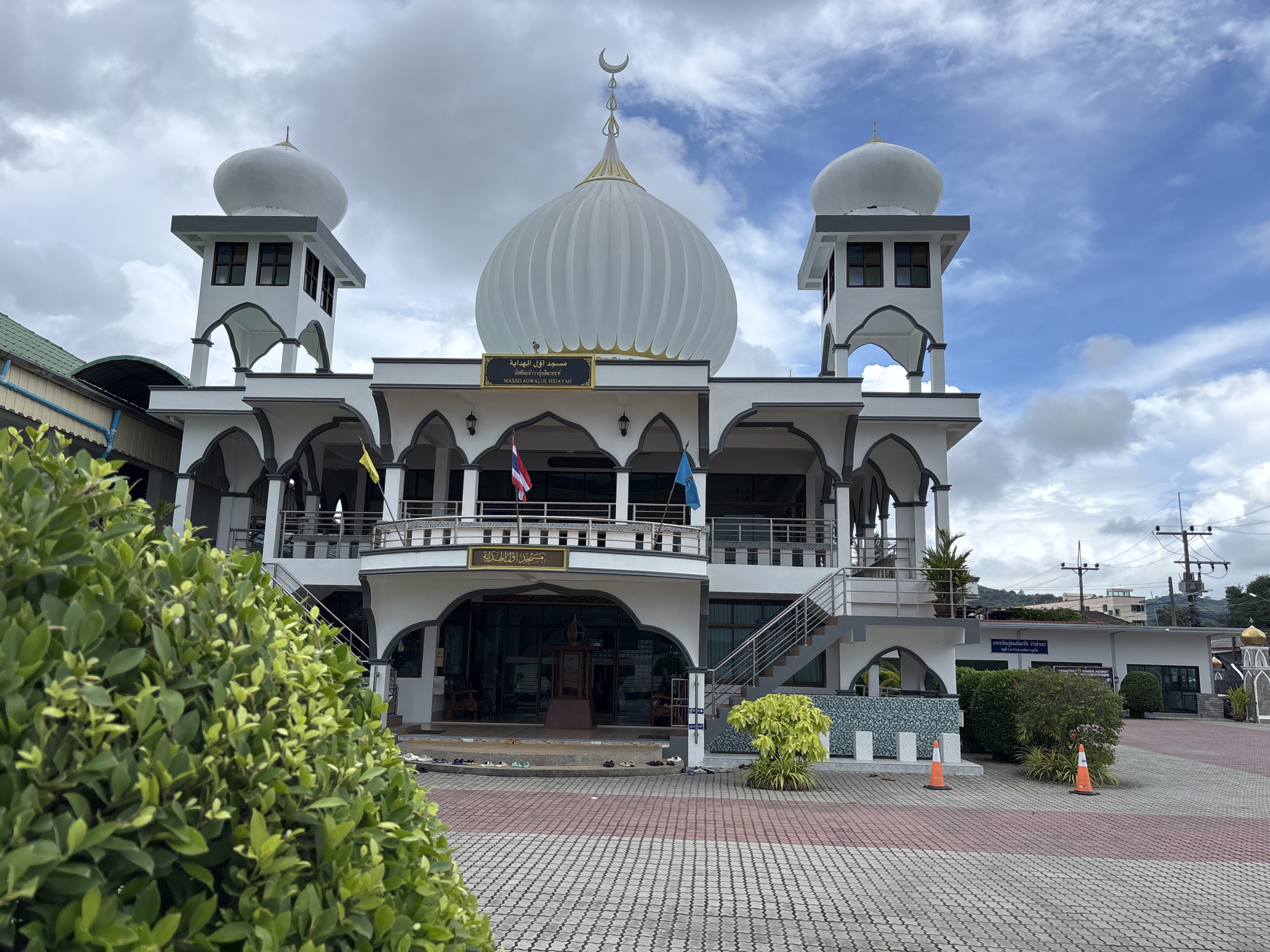 The image shows the Masjid Aowallul Hidayah in Rawai, Thailand, on a cloudy day. The mosque is a white building with a large central dome and two smaller domes on towers on either side. The mosque has a traditional Islamic architectural style with arched windows and doorways. The building is surrounded by a paved courtyard and some greenery. The entrance to the mosque is visible, with shoes placed outside. There are no people visible in the image.
