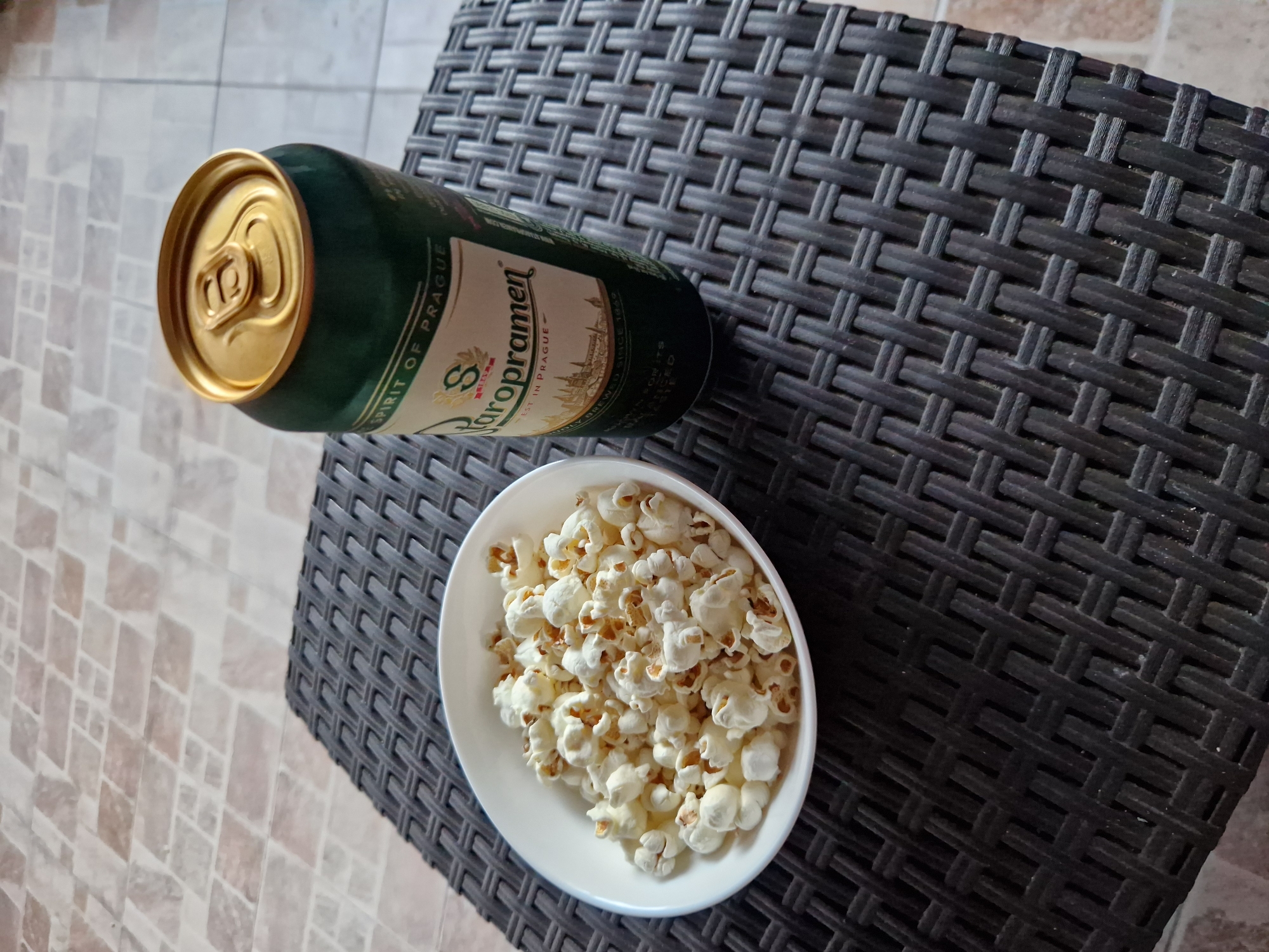 A close-up, high-angle shot shows a white bowl filled with popcorn and a green can of Staropramen beer with a gold pull-tab. Both items are on a dark, woven surface, which is resting on a tiled patio. The scene suggests a casual outdoor setting, potentially a backyard or balcony in Sofia, Bulgaria, given the recognizable Staropramen beer brand, which is brewed in the Czech Republic but widely available and popular in Bulgaria. The tiles on the patio appear to be a light beige and brown, laid in a staggered pattern.