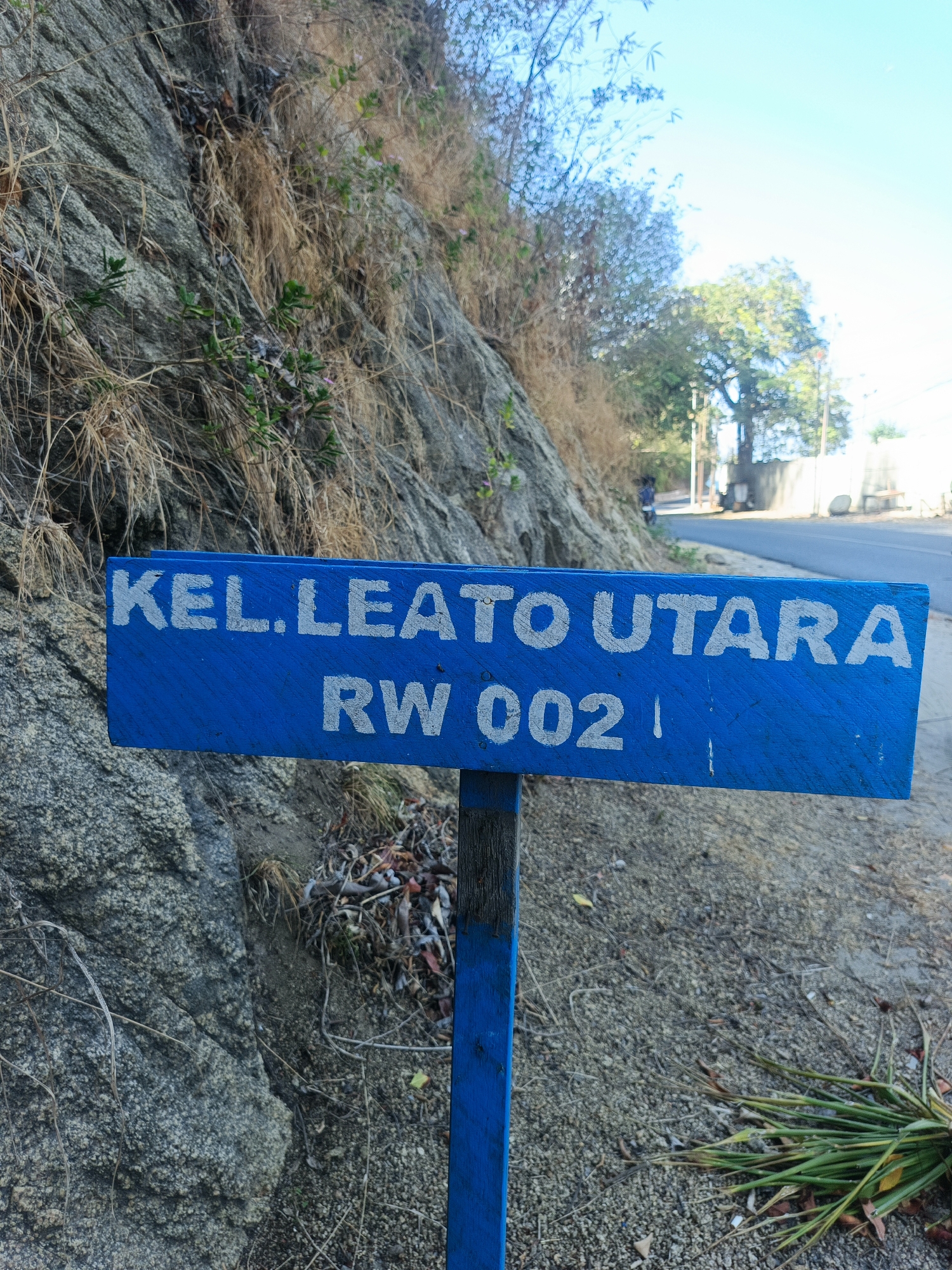 A blue wooden sign is partially visible in the foreground of an outdoor scene. The sign has white lettering and reads "KEL. LEATO UTARA" on the top line and "RW 002" on the bottom line. The sign is mounted on a blue wooden post. The background consists of a rocky hillside with dry grass and some green foliage. A paved road is visible in the distance, with a person walking on it and trees lining the side of the road. The sky is clear and blue. The scene appears to be in a semi-urban or rural setting in Gorontalo, Indonesia, given the presence of the "RW" designation, which stands for Rukun Warga (Neighborhood Association) in Indonesia.