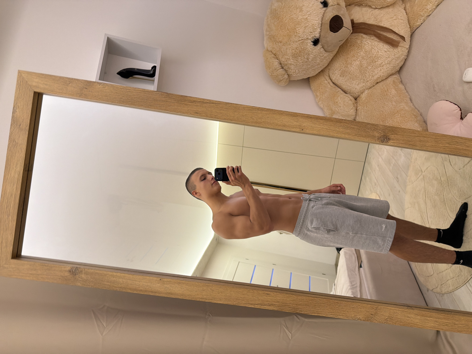 The image shows a young man taking a mirror selfie in what appears to be a bedroom. He is shirtless and wearing grey sweatshorts and black socks. He is holding a smartphone in his right hand. The mirror is tall and has a wooden frame. To the right of the mirror, a large teddy bear is visible, sitting on a bed. On the wall above the teddy bear, there is a small white shelf with a black high-heeled shoe on it. The room has light-colored walls and a tiled floor. A white door with blue horizontal stripes is visible in the background. The overall impression is that of a casual personal space. There is no identifiable information in the image that would indicate the location in Elhovo, Bulgaria.