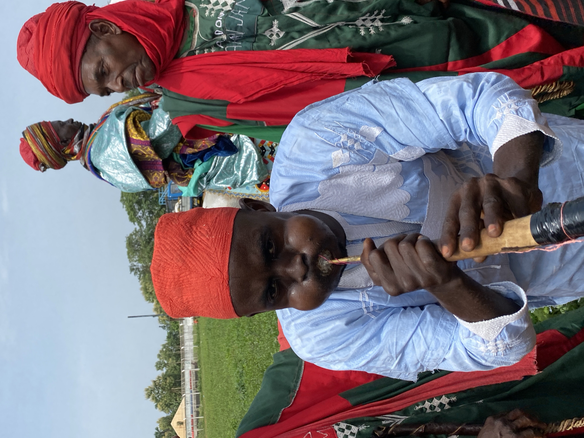 In Kafanchan A, Nigeria, a man in the foreground, wearing a red cap and light blue robe, plays a woodwind instrument. His cheeks are puffed out. Behind him are two more men in traditional attire, one with a red turban and the other with a multicolored headdress. All three are standing on a patterned cloth on a grassy field near a fence. In the background, there are trees and a light blue sky.