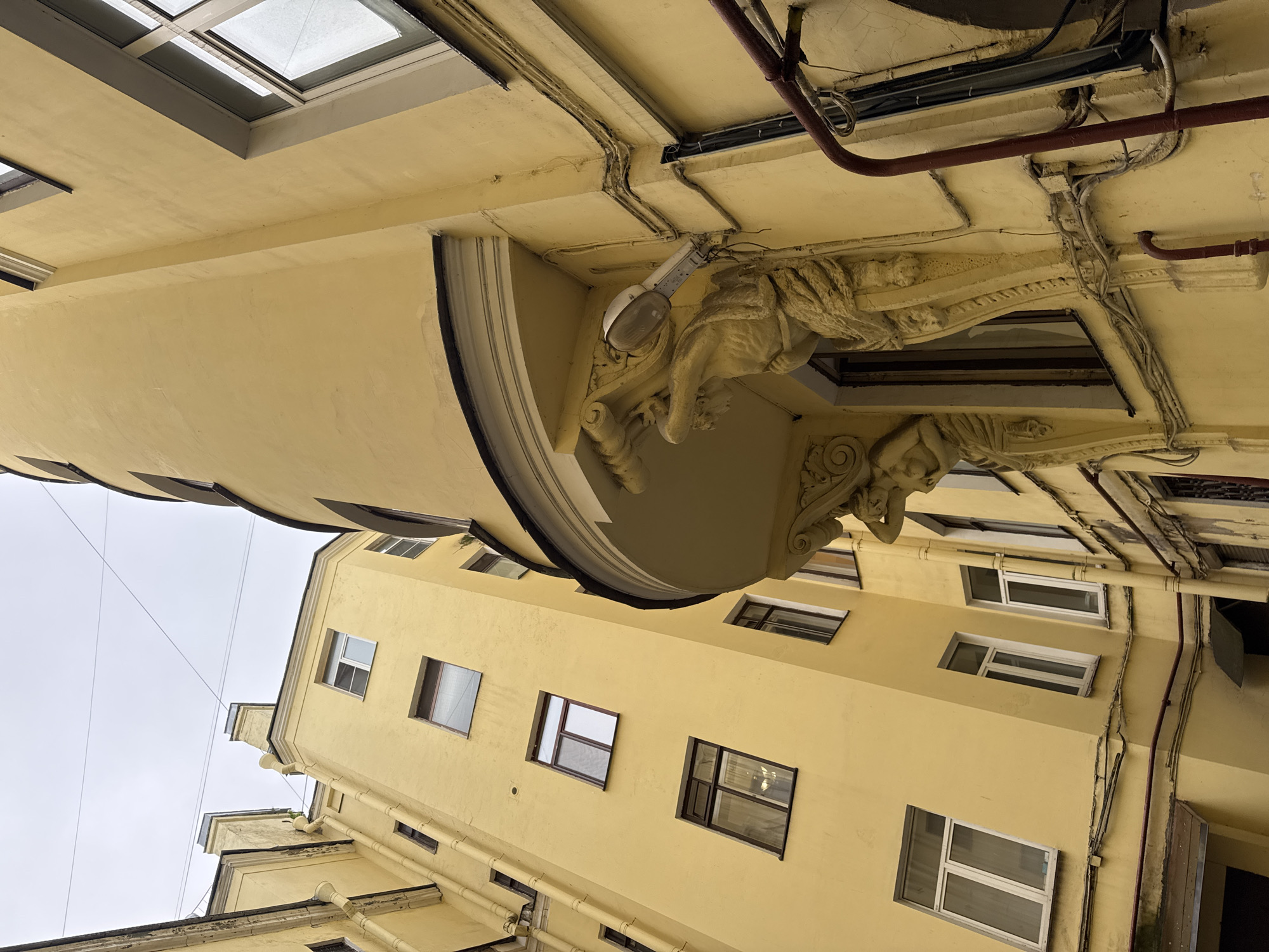 The image shows an upward view of the courtyard of a historic building in Saint Petersburg, Russia, likely in the central part of the city. The architecture is characterized by narrow, deep courtyards, a common feature of St. Petersburg's urban planning. The buildings are painted a pale yellow and feature classical architectural elements, such as ornate stucco work. In the foreground, the image captures a close-up of a balcony, supported by sculpted caryatids, which are female figures used as architectural supports. A modern streetlight is affixed to the building near the balcony, and electrical conduits run along the facade. Multiple windows of various sizes and designs are visible on the building's exterior. The sky above is overcast and pale, typical of the city's climate. The perspective emphasizes the height of the buildings and the confined nature of the courtyard.