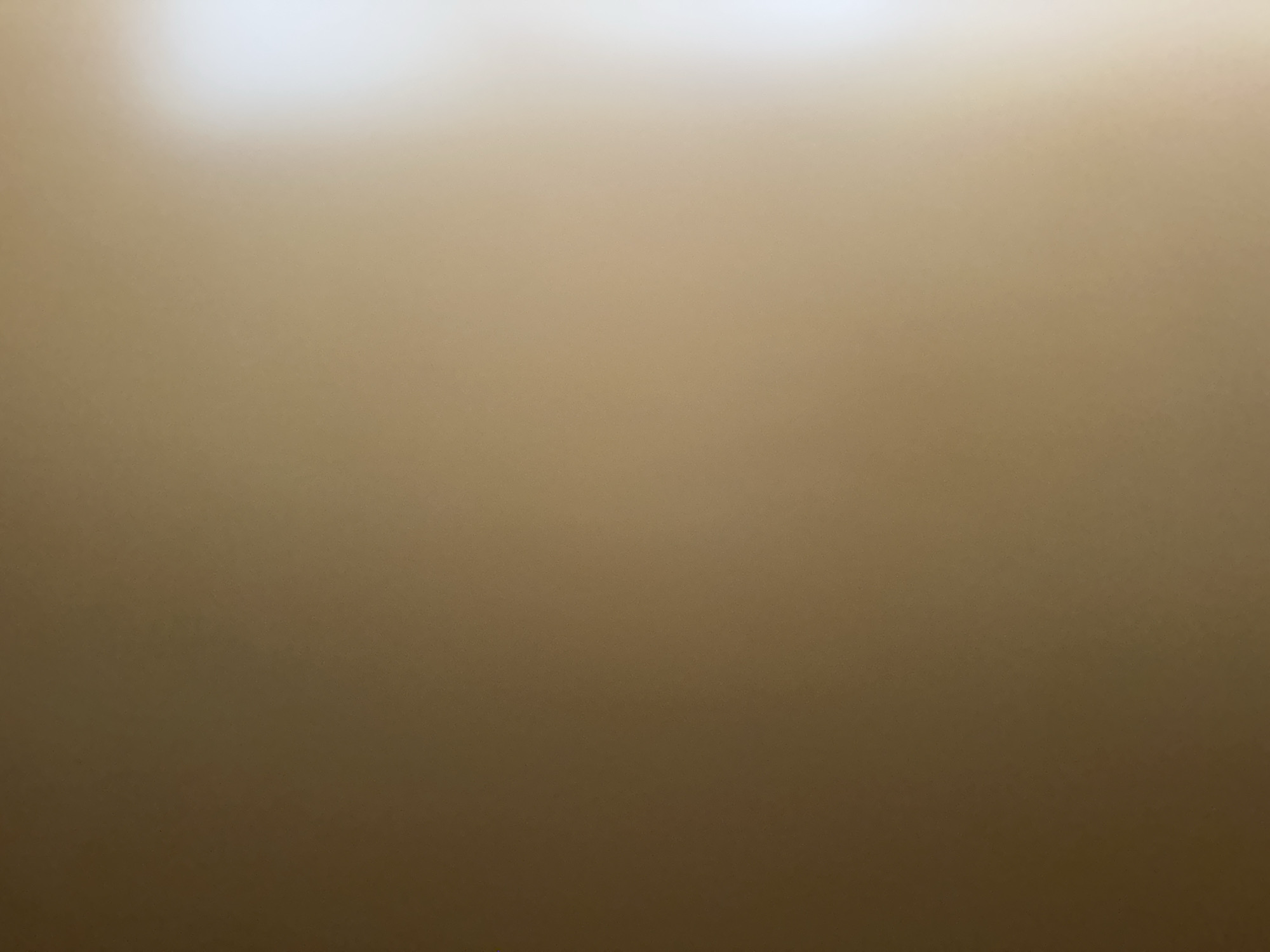 The image is a close-up, out-of-focus shot of a wall with a gradient of light brown to a lighter, almost white hue at the top. There are no discernible subjects, objects, or activities taking place. The lighting suggests a bright light source above and to the left, possibly an overhead light or sunlight from a window. There is no visible text. The scene is abstract due to the lack of focus and detail. Given the provided location context of Belgrade, Serbia, and the nature of the image, it's possible this is a detail of an interior wall in a building, but without any clear visual cues, this is purely speculative. The overall impression is one of muted tones and a lack of defining characteristics, making it difficult to ascertain any specific context or meaning.