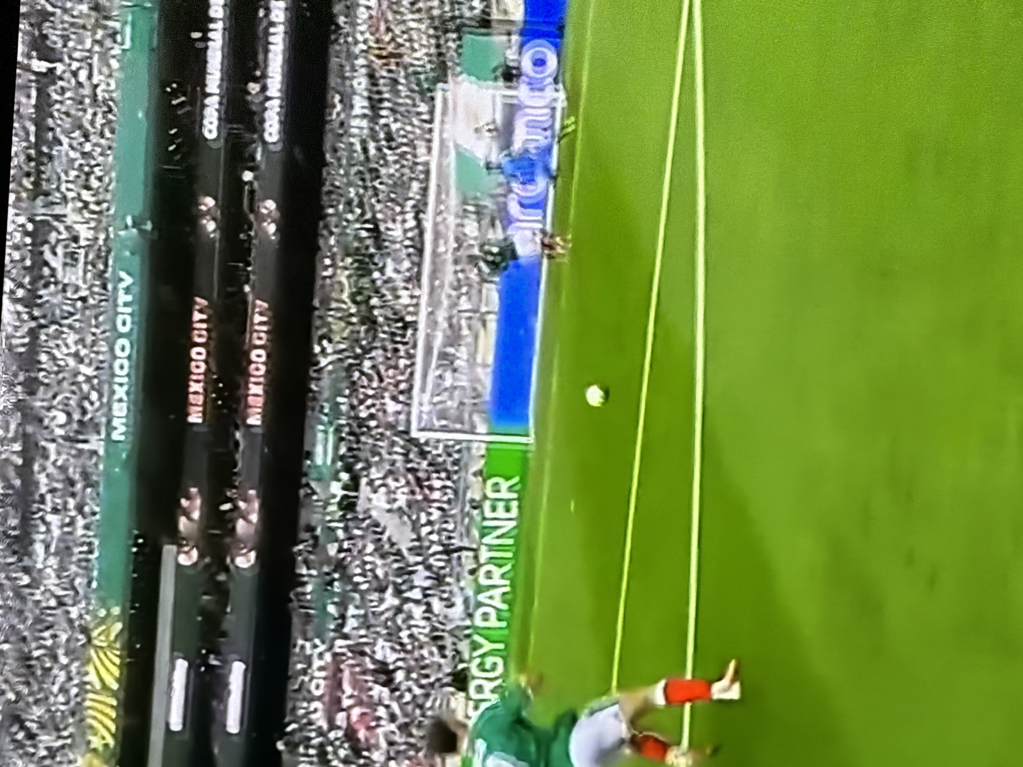 The image captures a soccer match in a stadium. In the foreground, a soccer player in a green jersey and white shorts with red accents is kicking a soccer ball on a vibrant green field. The player's left foot is on the ball, and their right leg is extended. In the background, a goal net is visible, with other players in blue jerseys behind it. The stands are filled with spectators, and prominent advertising boards display "MEXICO CITY" and "COPA MUNDIAL DE". The scene suggests an active sporting event taking place in a large stadium environment.
