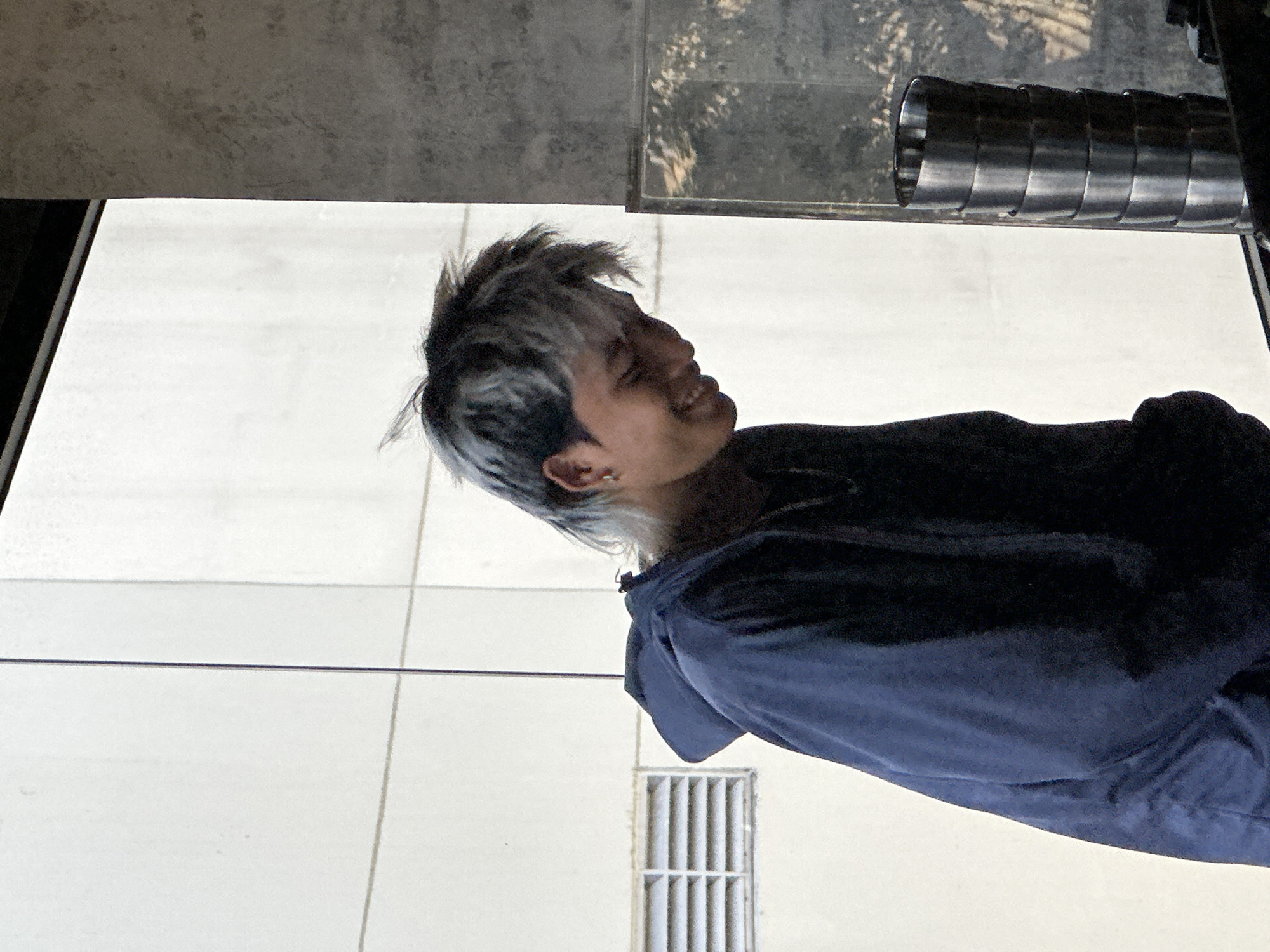The image depicts a young man with styled, multi-colored hair, wearing a dark blue hooded jacket. He is looking down and smiling, with his right ear visible, adorned with a small hoop earring. He is positioned in what appears to be an indoor setting, possibly near a large window or partition, with a textured gray wall to his right and a white, tiled or paneled wall to his left. A stack of metallic cylindrical objects, resembling tumblers or cups, is visible on the right side of the frame. The environment suggests an urban setting, possibly a café or a commercial space in Đà Nẵng, Vietnam. The lighting is somewhat diffuse, creating soft shadows.