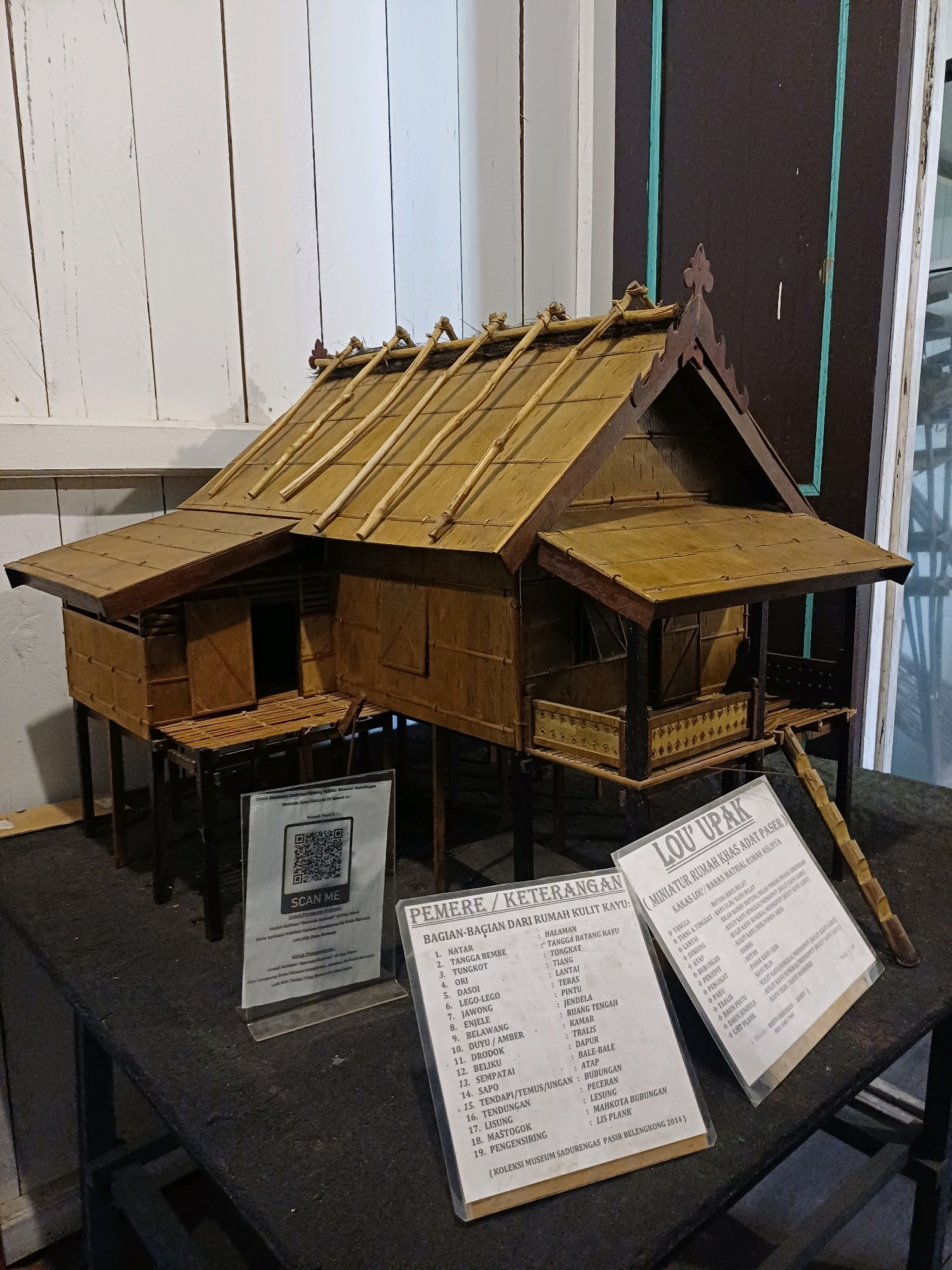 The image depicts a model of a traditional house, specifically identified as a "Rumah Adat Pasir Belengkong" or "Rumah LOU EPAK," situated in the city of Pasir Belengkong, Indonesia. The model is constructed from natural materials such as bamboo and wood, showcasing the architectural style with a raised structure supported by stilts, a sloping roof thatched with bamboo, and intricate decorative elements on the walls and railings.
In front of the model, several informational displays are placed on a dark, textured surface, likely a table or stand. One display, with a QR code and the text "SCAN ME," is positioned directly in front of the house model. To the right of this, another sign titled "LOU EPAK (MINIATUR RUMAH KHAS ADAT PASER)" provides further details about the house, including its materials and components. To the left of the QR code display, a sign labeled "PEMERE / KETERANGAN BAGIAN-BAGIAN DARI RUMAH KULIT KAYU" lists and describes various parts of the house.
The background consists of vertical wooden panels on the left and a darker, possibly wooden, panel with vertical teal stripes on the right. The lighting appears to be artificial, casting shadows and highlighting the textures of the model and the displays. The overall scene suggests an indoor exhibition or museum setting, dedicated to showcasing local heritage and architecture. There are no people visible in the image.