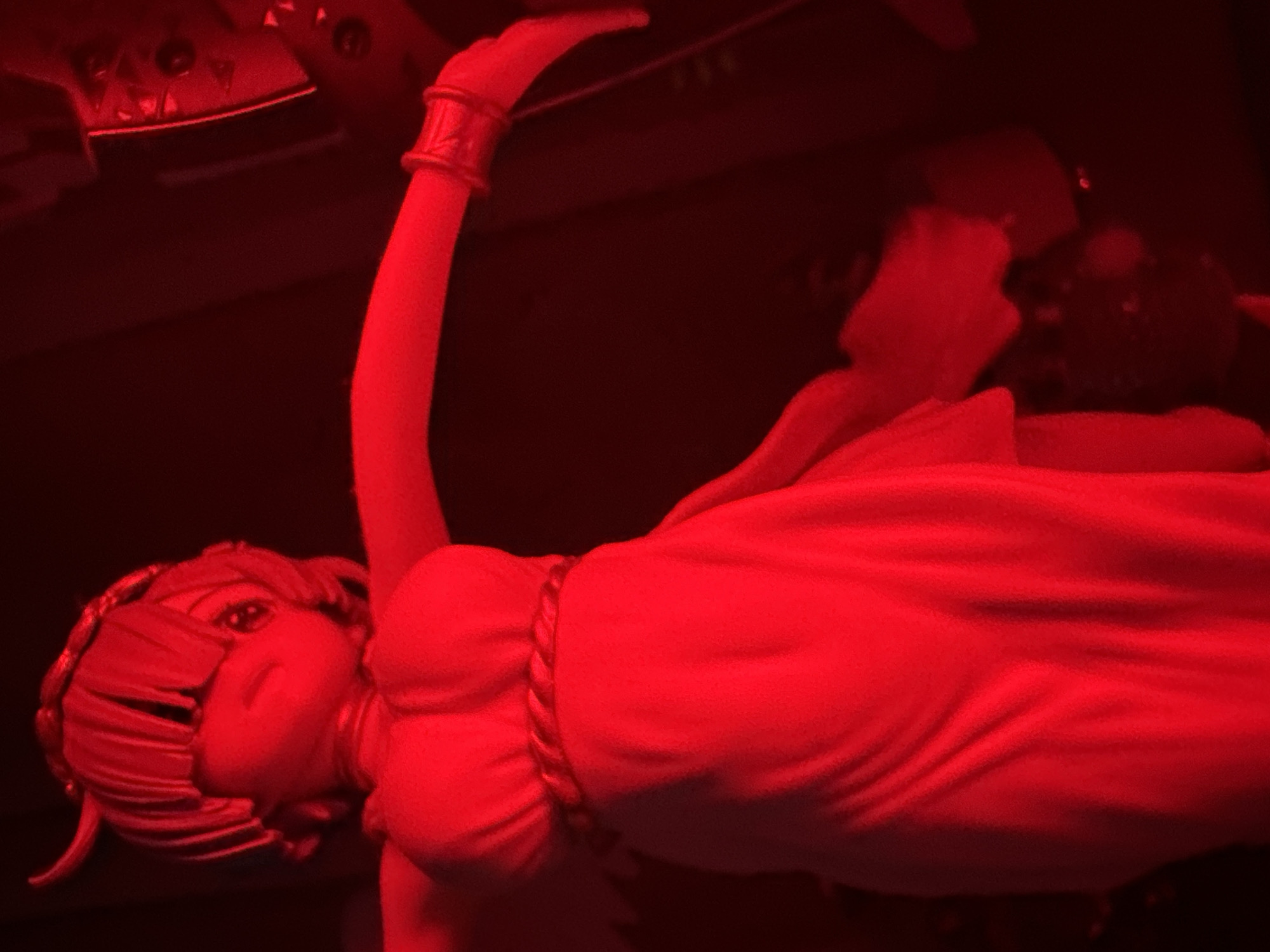 An anime-style figurine is depicted in a dimly lit environment, bathed in red light. The figurine appears to be a female character with short, straight hair and large eyes. She is wearing a strapless white dress with a braided rope detail around her waist. Her arms are outstretched, and she has a bracelet on her right wrist. The background is dark and out of focus, with some indistinct shapes that might be other objects or decorations. The red lighting casts strong shadows and gives the scene an atmospheric, almost dramatic quality.