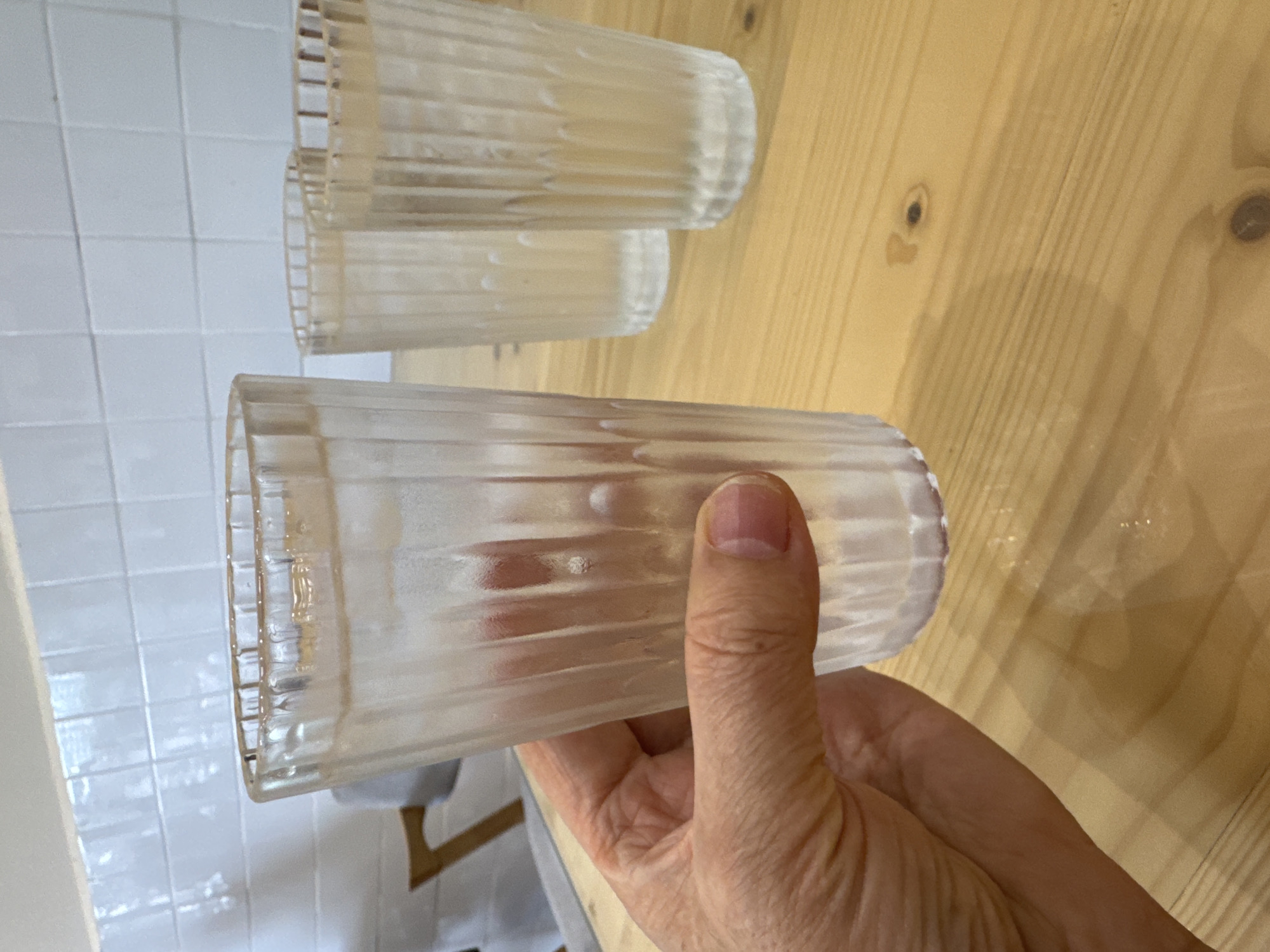 A close-up shot shows a hand holding a tall, fluted glass filled with ice and a clear liquid, likely water. Two other identical glasses are visible behind it, also on a light-colored wooden surface. The background features a white tiled wall. The overall scene suggests a casual setting, possibly a cafe or a home. There is no discernible location in Kuala Lumpur, Malaysia.