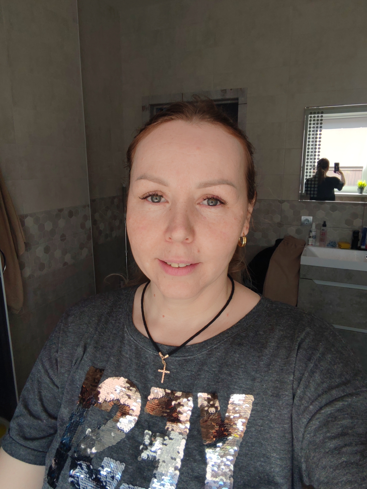 A woman is taking a selfie in a bathroom. She has red hair and green eyes and is wearing a gray t-shirt with sequins and a gold cross necklace. She is smiling slightly. The bathroom has gray walls and a mirror. A second person is visible in the mirror, taking a photo outside a window with their back to the bathroom. The location is Chaikovsky, Russia.