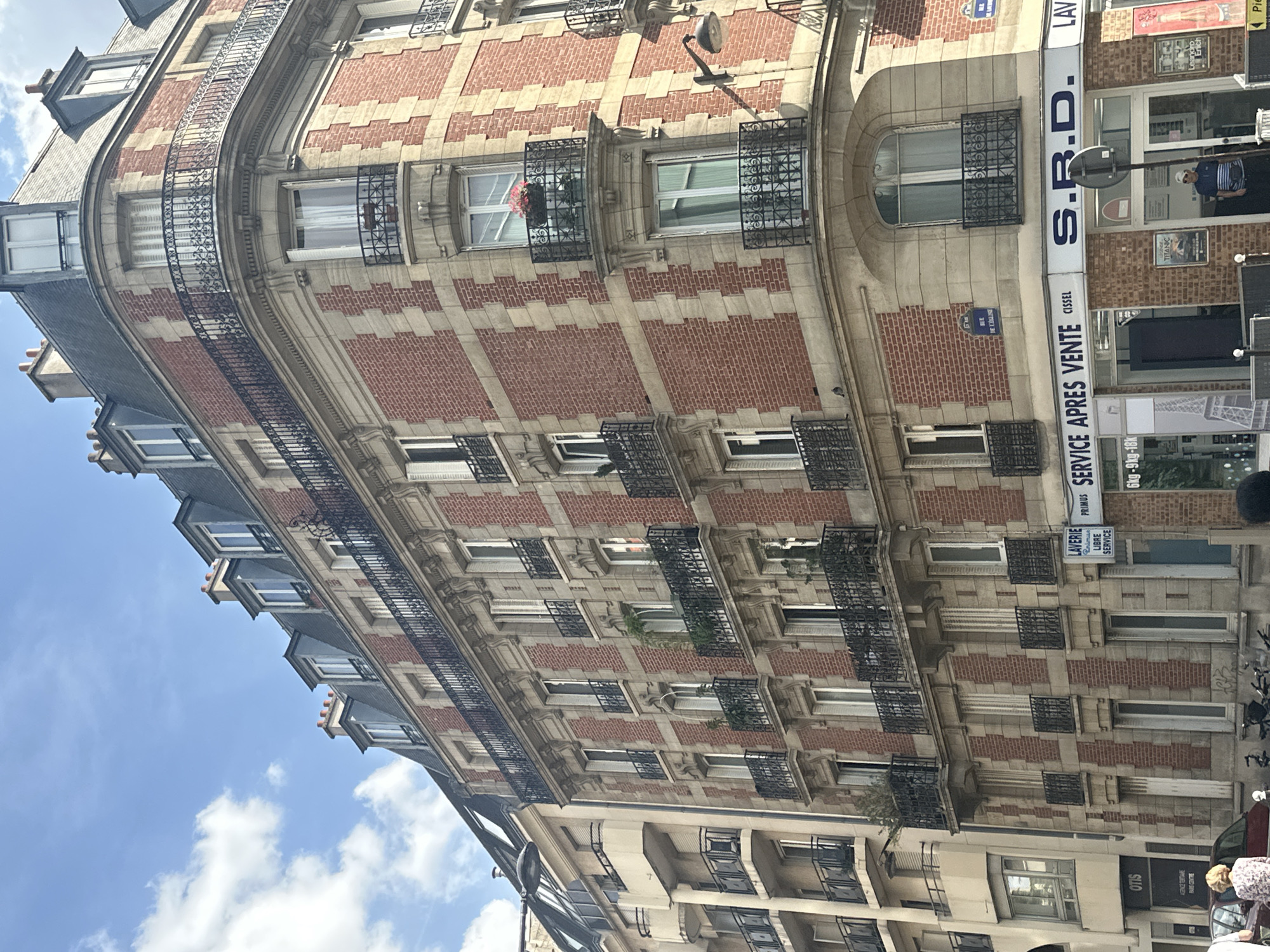 The image shows a street scene in Paris, France, focusing on the corner of a multi-story brick building with stone accents. The building features multiple balconies with ornate black railings, mansard roofs with dormer windows, and several windows with white shutters. Some balconies have plants. On the ground floor, a business with signage that includes "S.R.D." and "LAVERIE SERVICE APRES VENTE" is visible. The street appears to be moderately busy, with a few people visible on the sidewalk and a car parked at the curb. The sky above is blue with scattered white clouds. Another building with a lighter facade and balconies is situated to the left of the main building. A street sign with "RUE DE L'ÉGLISE" is attached to the corner building.