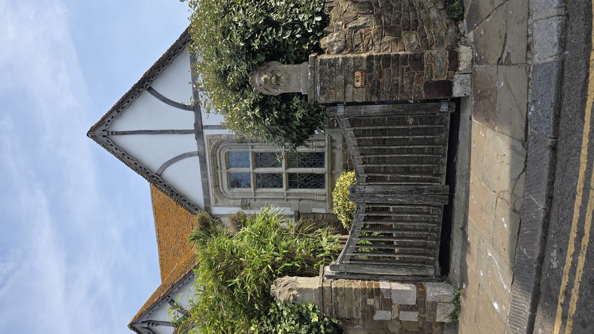 The image shows a picturesque scene in the city of St Leonards, United Kingdom, featuring a charming, old-fashioned house with a distinctive Tudor-style architectural design. The house has white plastered walls and exposed dark timber beams, with a steeply pitched, orange-tiled roof. A prominent feature is the ornate stone window, divided into multiple panes with leaded glass. In front of the house, a rustic wooden gate with vertical slats is flanked by stone pillars topped with carved lion heads. The gate is set within a low stone wall, and a "NO PARKING" sign is visible on the right-hand pillar. Lush green bushes and foliage frame the gate and the house, adding to the inviting atmosphere. The foreground shows a paved walkway with stone slabs, bordered by a street with yellow double lines painted on the asphalt. The sky above is a clear blue with a few scattered white clouds, suggesting fair weather. The scene appears tranquil and well-maintained, conveying a sense of historical charm and residential serenity.