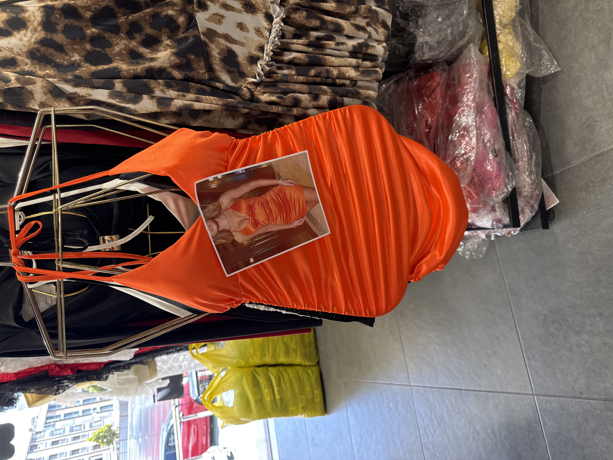 The image shows an orange ruched dress displayed on a clothing rack in a store. A photograph of a woman wearing a similar dress is pinned to the front of the displayed dress. Behind the orange dress, other clothing items are visible, including a leopard print dress and black garments. In the background, two yellow plastic bags are stacked on the floor, and a red car is seen through a window. The floor is tiled with grey square tiles. This scene is located in the city of Gungoren, Turkey.