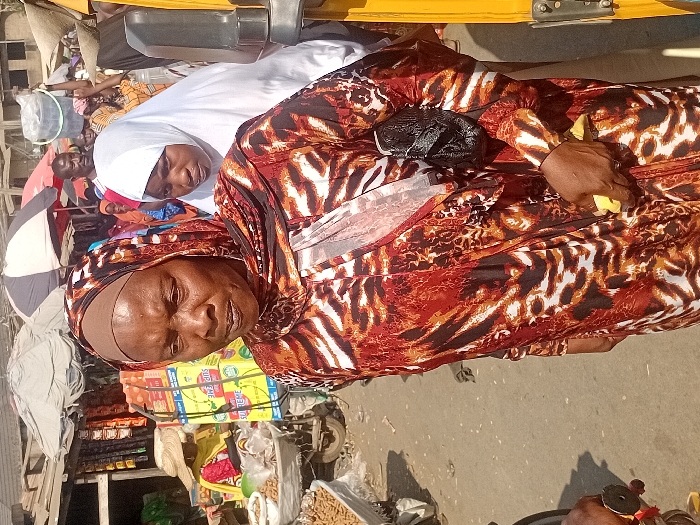 Two women are prominently displayed in the foreground. The woman in front wears a patterned headscarf and dress, with a purse on her chest. The other woman, behind and slightly to her left, is dressed in a white hijab. In the background, other people are visible amidst a market scene. Goods such as packaged foods are stacked on a cart or stand, hinting at a bustling commercial environment. A vehicle's frame is visible at the top of the image.