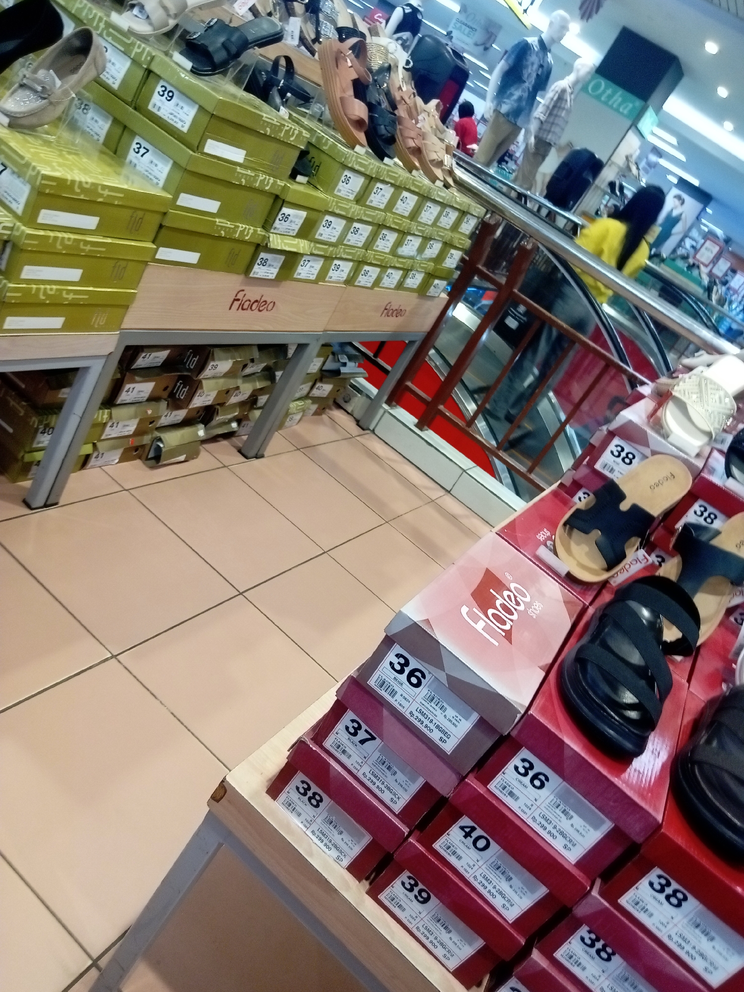 The image shows a retail store display of Fladeo brand footwear, specifically women's sandals, arranged on tiered shelving and stacked boxes. The setting appears to be an indoor shopping mall or department store, with tiled flooring, metal shelving units, and a glimpse of an escalator in the background. The lighting is bright and appears to be overhead fluorescent lighting typical of retail environments.
Multiple stacks of shoe boxes are visible, each labeled with brand information and size numbers, such as "36," "37," "38," "39," and "40." Some of the boxes are open, revealing sandals inside. Several pairs of sandals are also displayed on top of the boxes, showcasing various designs, predominantly black and tan colors. The brand name "Fladeo" is prominently displayed on the boxes and in some of the sandal designs.
There are no visible people in the immediate foreground or interacting with the products. However, in the background, the blurred figures of shoppers are seen near an escalator, suggesting an active retail environment. The overall impression is a well-organized display of footwear in a commercial setting. Based on visual cues such as store branding and product offerings, this scene is consistent with retail environments commonly found in shopping centers in Batam, Indonesia, which is known for its various malls and department stores.
