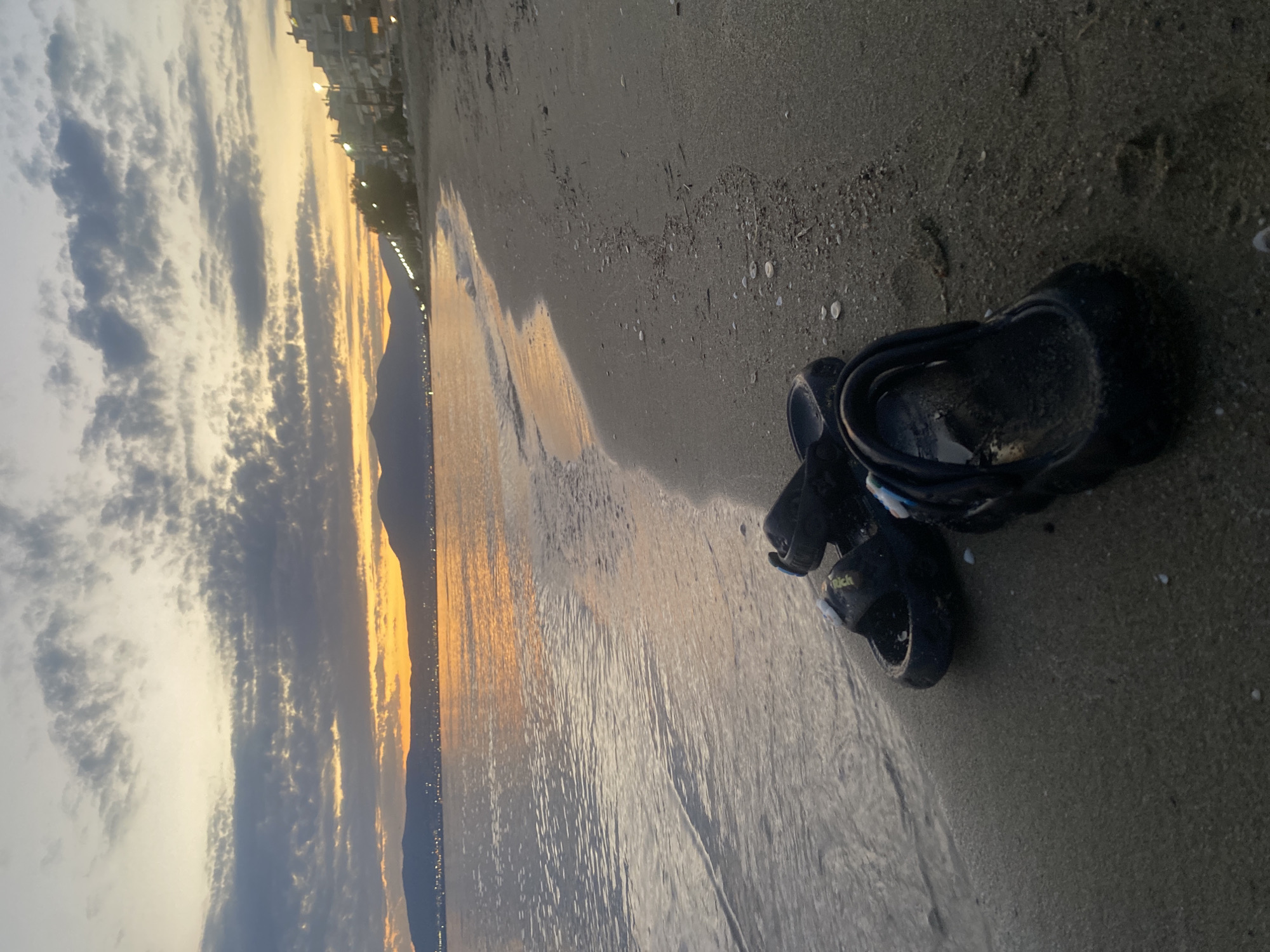 The image depicts a beach scene at dusk in Neoi Epivates, Greece. Dark-colored sandals are resting on the sandy shore, with the gentle waves of the sea lapping at the edge of the beach. In the background, a distant shoreline is visible with buildings and lights, and mountains form a silhouette against the twilight sky. The sky itself is painted with hues of orange, yellow, and gray clouds, creating a serene sunset atmosphere. The lighting is dim, consistent with the evening hour.