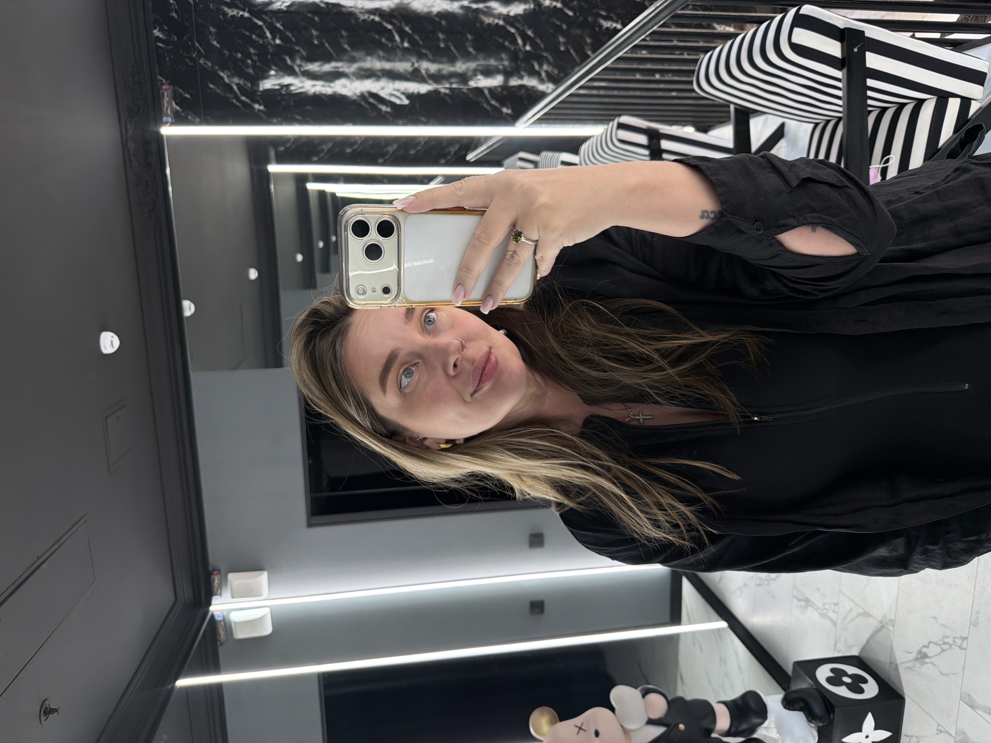A woman takes a mirror selfie in a modern, well-lit interior, likely a retail space, dressing room, or upscale bathroom in Moscow, Russia. She holds a gold-cased smartphone in her left hand, which has a ring with a green stone on her ring finger and light pink nail polish. Her long, light brown hair with highlights frames her face, and she has blue eyes and a neutral expression, looking directly into the phone's camera reflection. She is wearing a black long-sleeved top or dress, and a small tattoo, possibly reading "נשמה" (Hebrew for "soul"), is visible on her inner left forearm.
The setting features dark grey walls and a distinctive black and white marbled or patterned surface, possibly on the ceiling or an accent wall. Bright, horizontal LED light strips illuminate the space. In the background, visible in the mirror's reflection, are black and white striped cushions or upholstery, suggesting stylish furniture. On the floor, near the bottom right, is a black cube adorned with white, four-petal floral logos reminiscent of Louis Vuitton. Resting on this cube is a small, KAWS-style art toy or figurine with a white head, pink nose, and black and white body parts. Small white square dishes are visible on a reflective surface or shelf in the reflections. The overall ambiance is contemporary and chic.